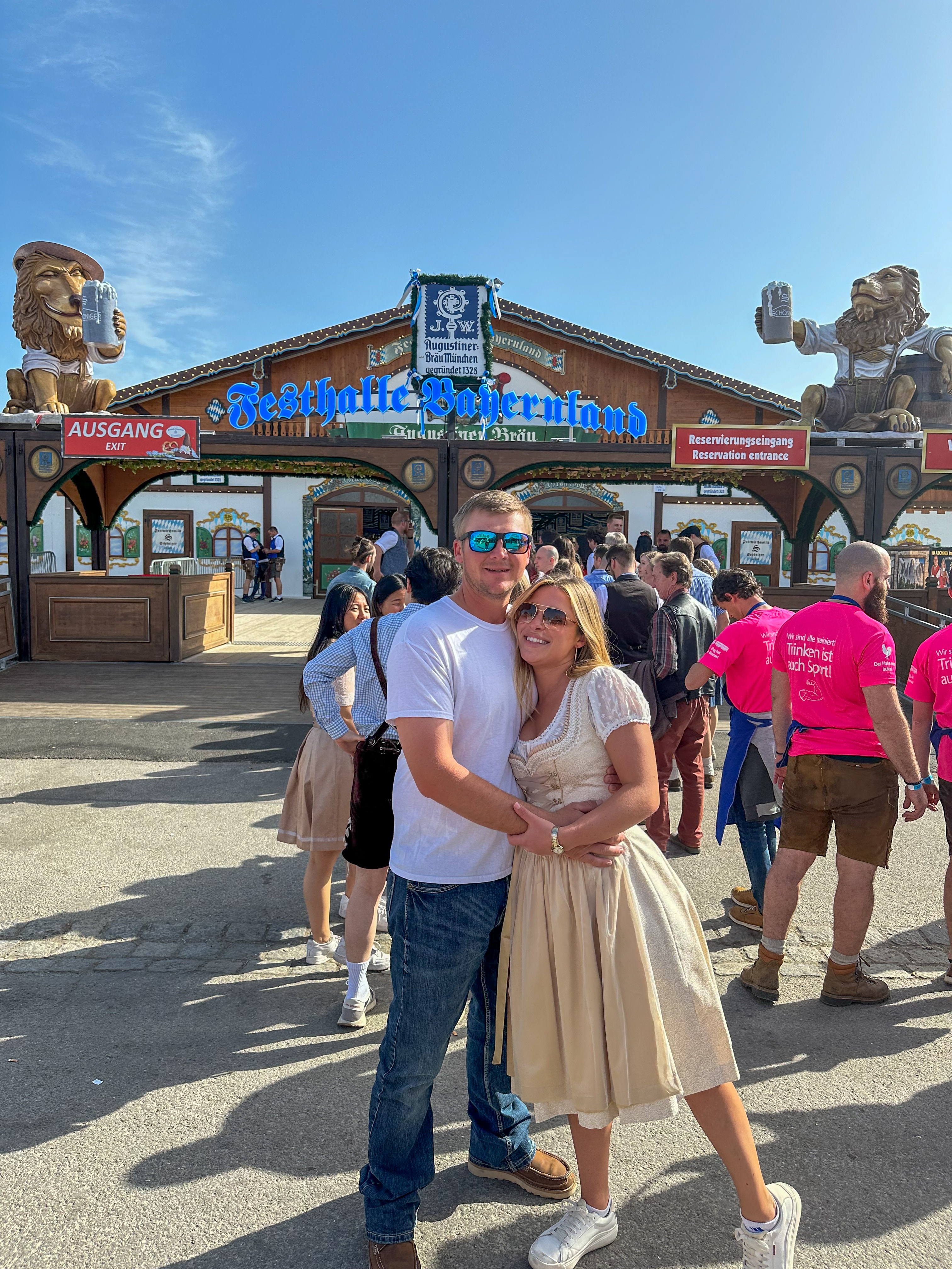 Hannah in a beige dress with her arm around a man in front of the entrance to an Oktoberfest event on a sunny day