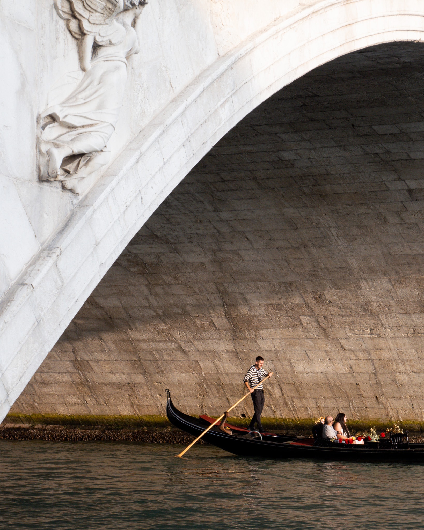 View of a two people riding in a gondola under a large bridge in Venice