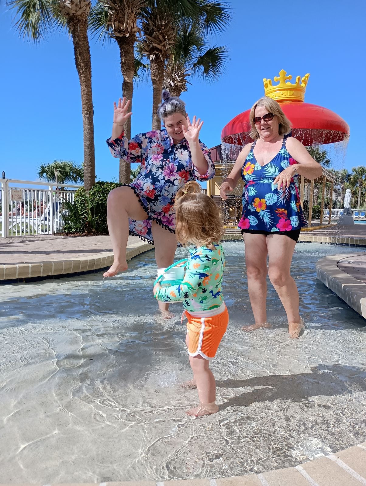 Two women in colorful shirts playing in shallow water with a toddler at a resort pool