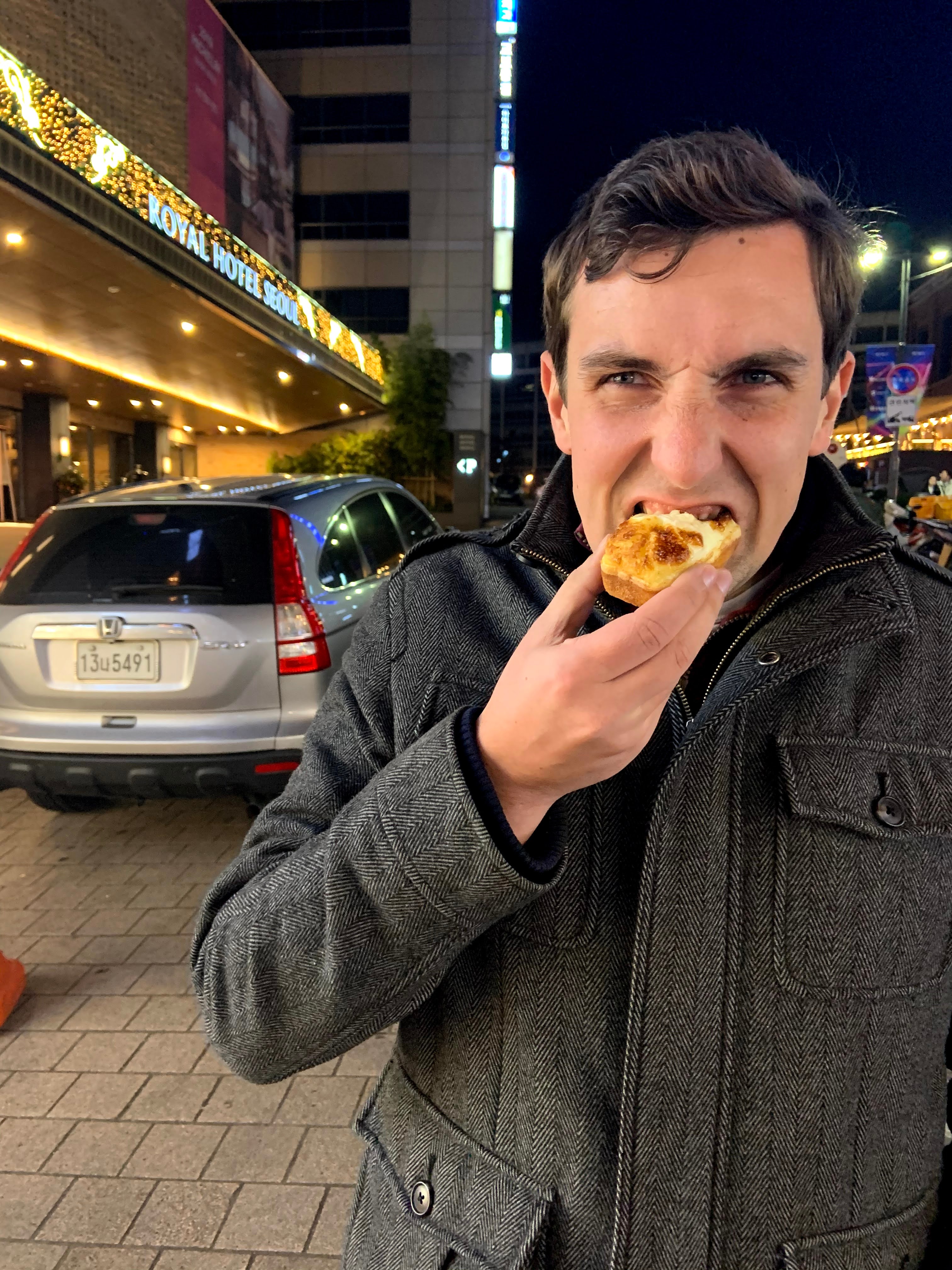 Image of a man in a grey coat biting a pastry on a city street at night