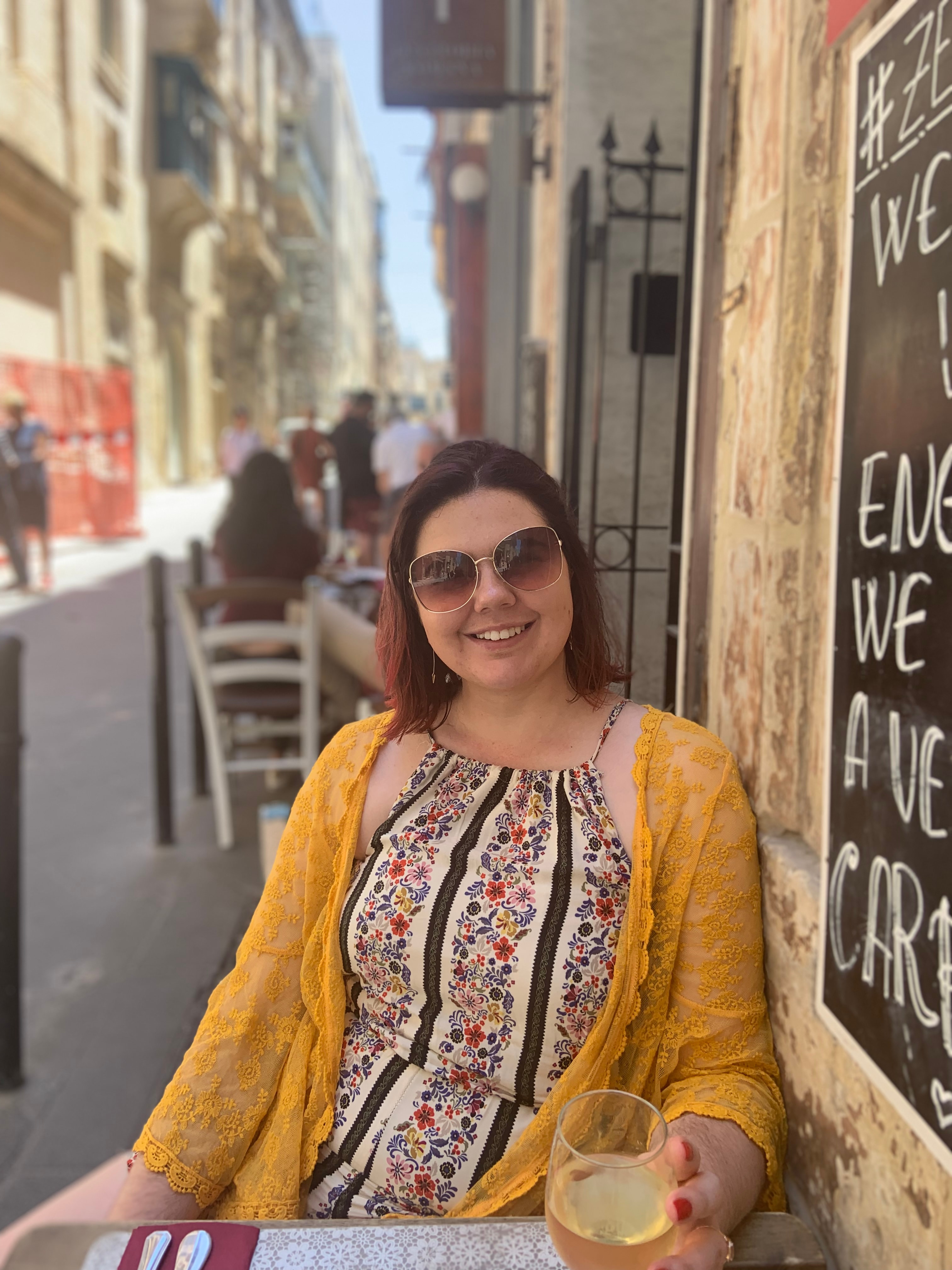 Ashley in a yellow cardigan and sunglasses sitting at an outdoor table with a glass of white wine