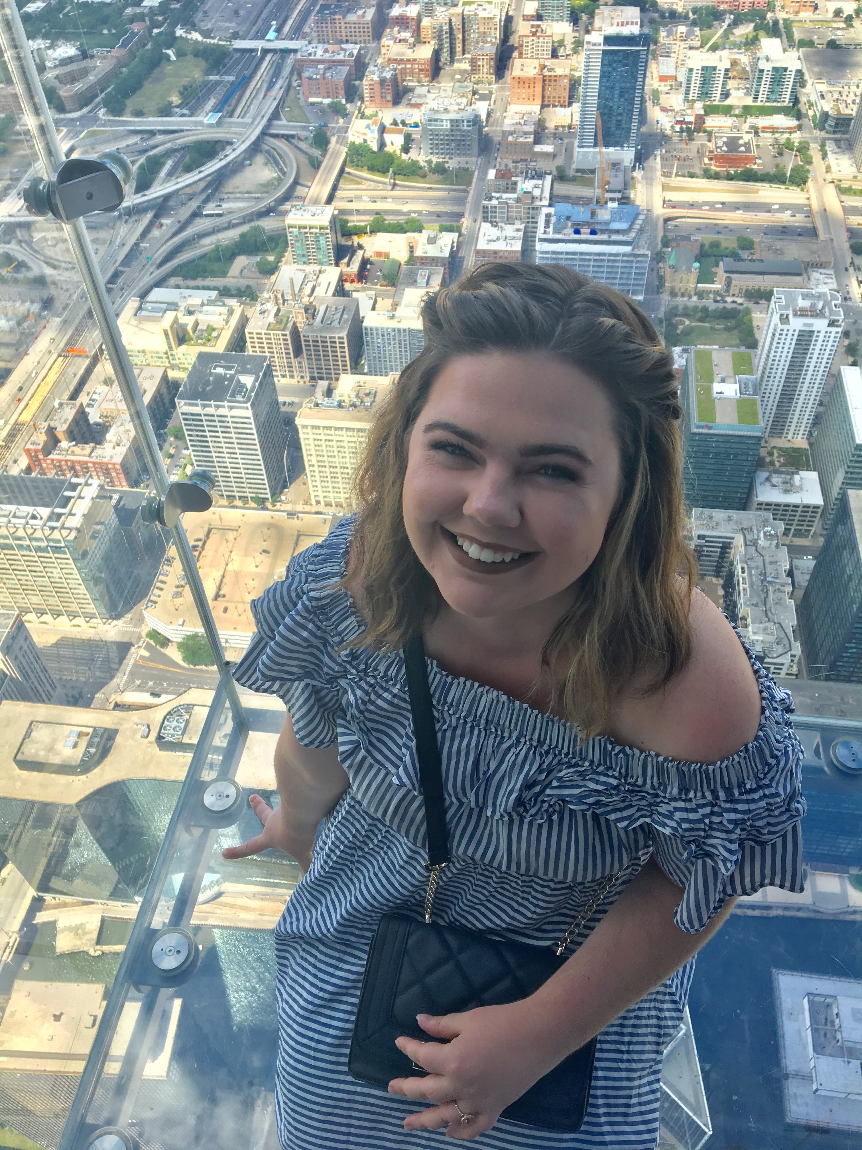 Ashley in a blue and white striped dress sitting on a glass balcony with views of a city below