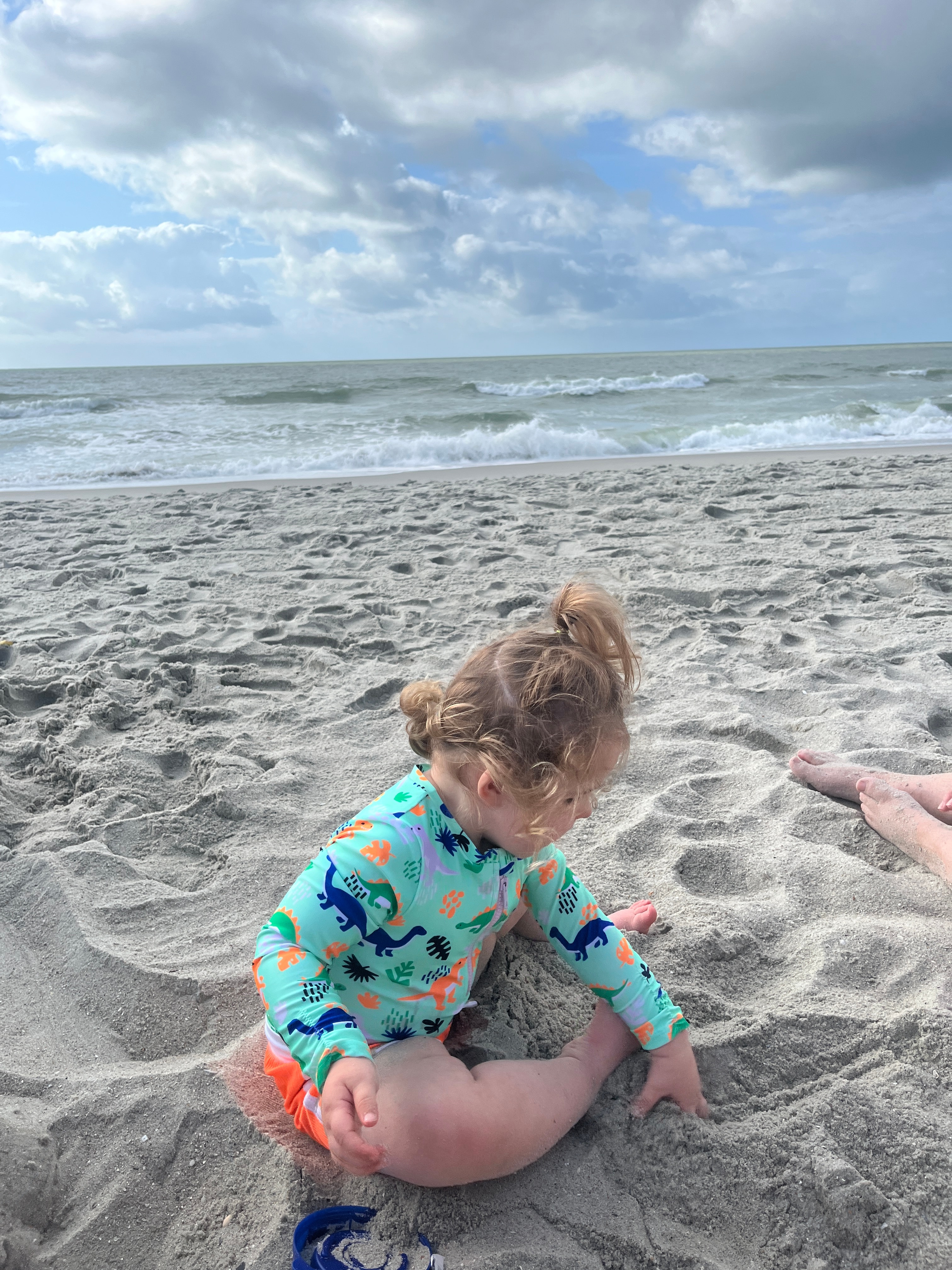 Image of a baby in a blue shirt playing in the sand on the beach with clouds above