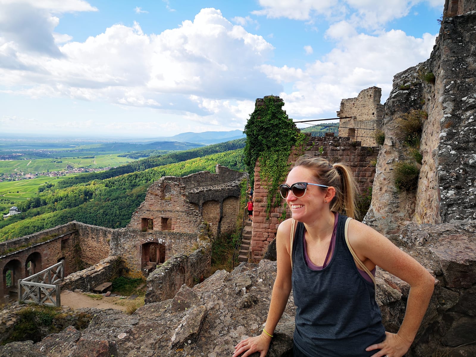 Amanda wearing sunglasses and smiling by old stone buildings with a pretty green valley view behind her