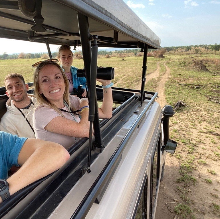 Amanda holding a camera with others in a safari vehicle driving through a field on a sunny day