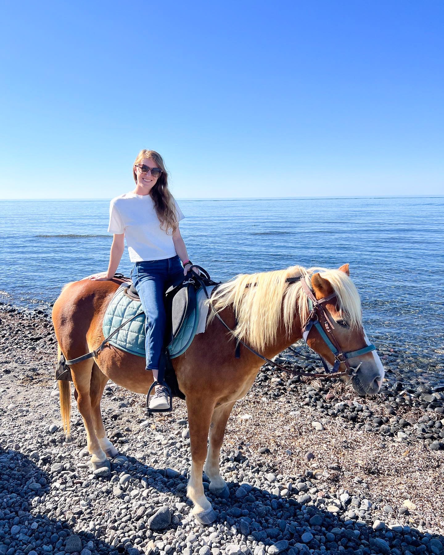 Kayla in jeans and a white t-shirt on horseback on a trail overlooking the ocean under clear skies