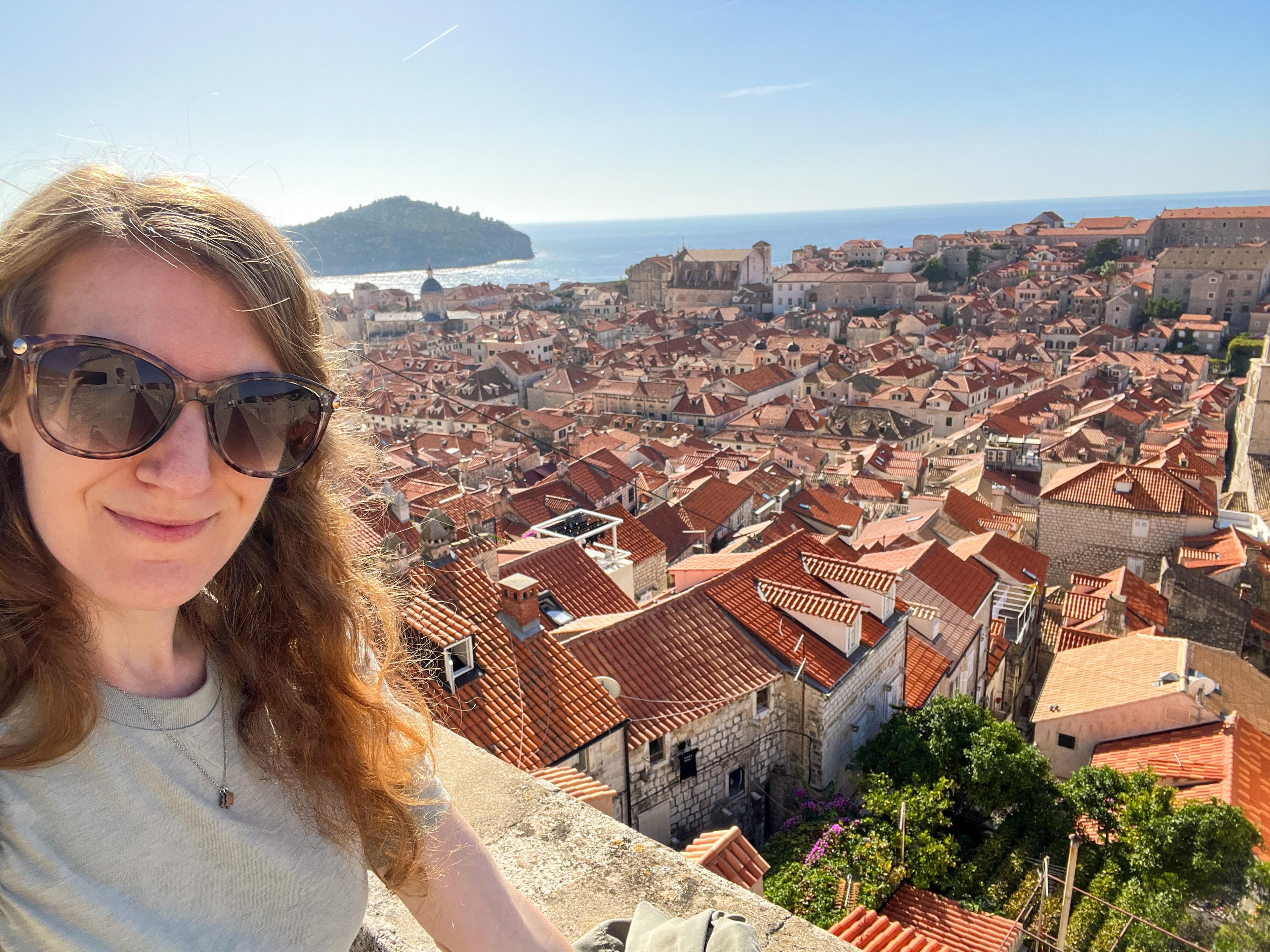 Kayla in sunglasses taking a selfie from a viewpoint overlooking orange rooftops in a coastal town, the ocean visible behind her