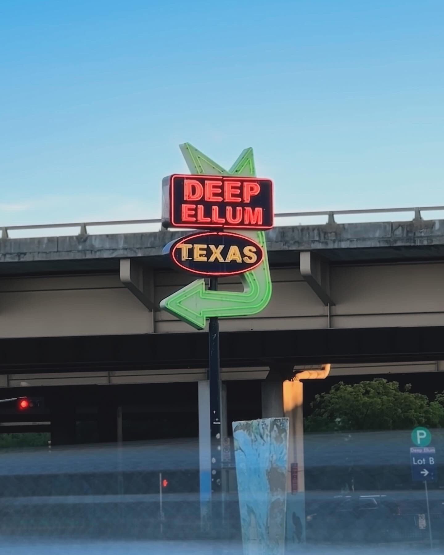 View of a neon sign hung from an overpass reading “deep ellum, Texas” with a green arrow pointing to the left