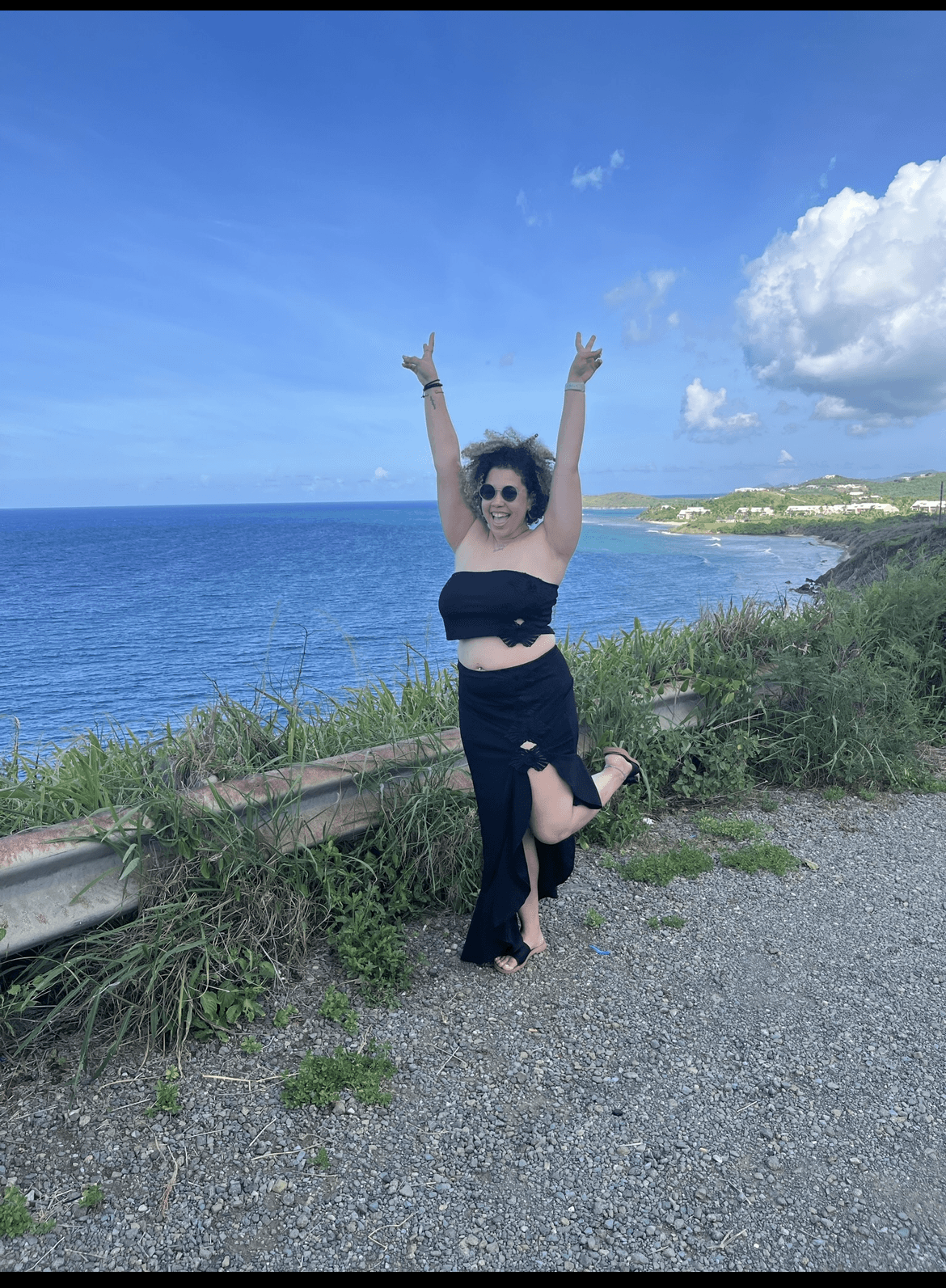 Bethany in a black skirt and matching top with arms raised to the sky and an ocean view behind her