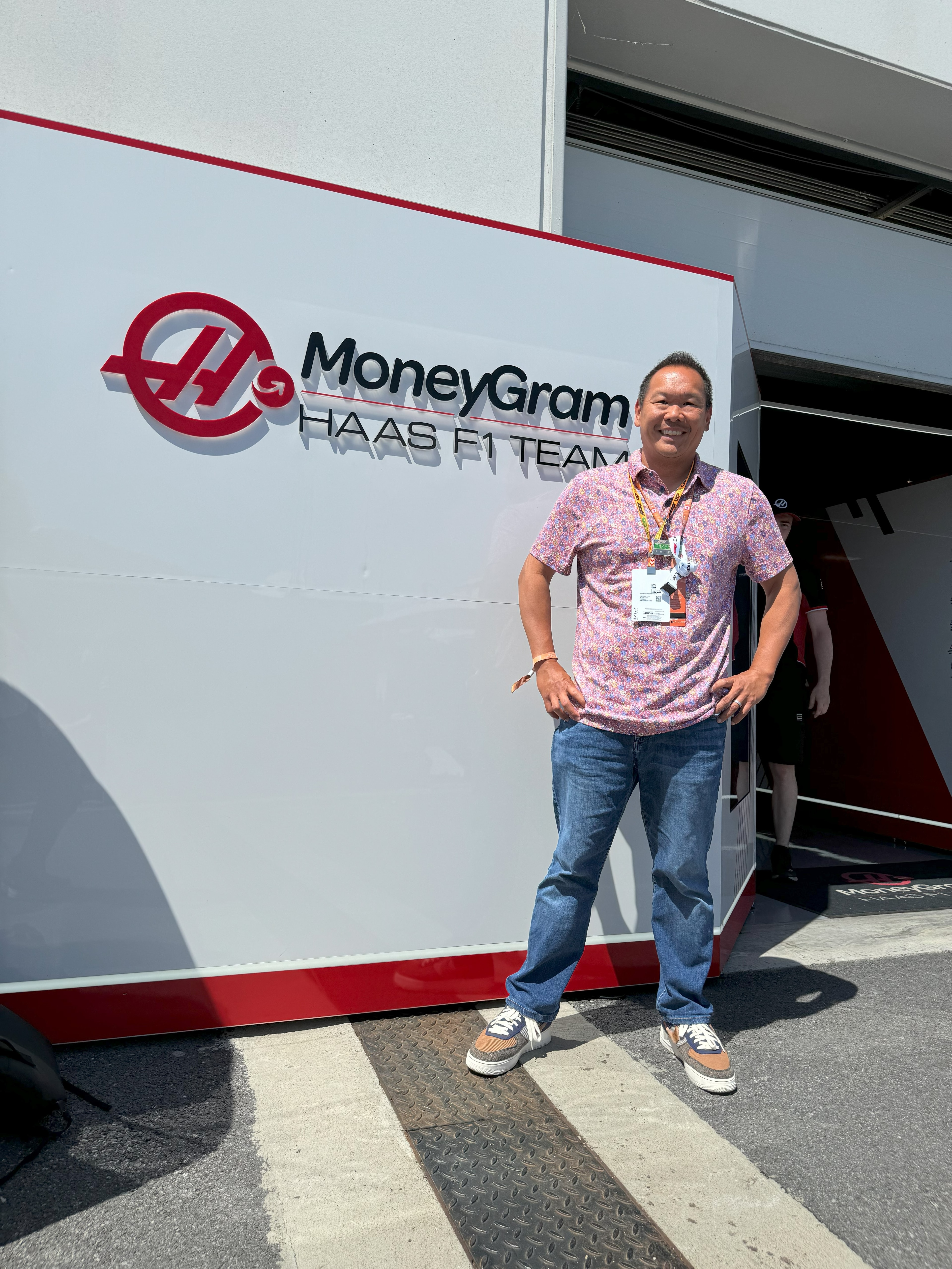 Robert in a pink shirt and jeans standing in front of a white wall with a sign reading “MONEYGRAM” outdoors on a sunny day