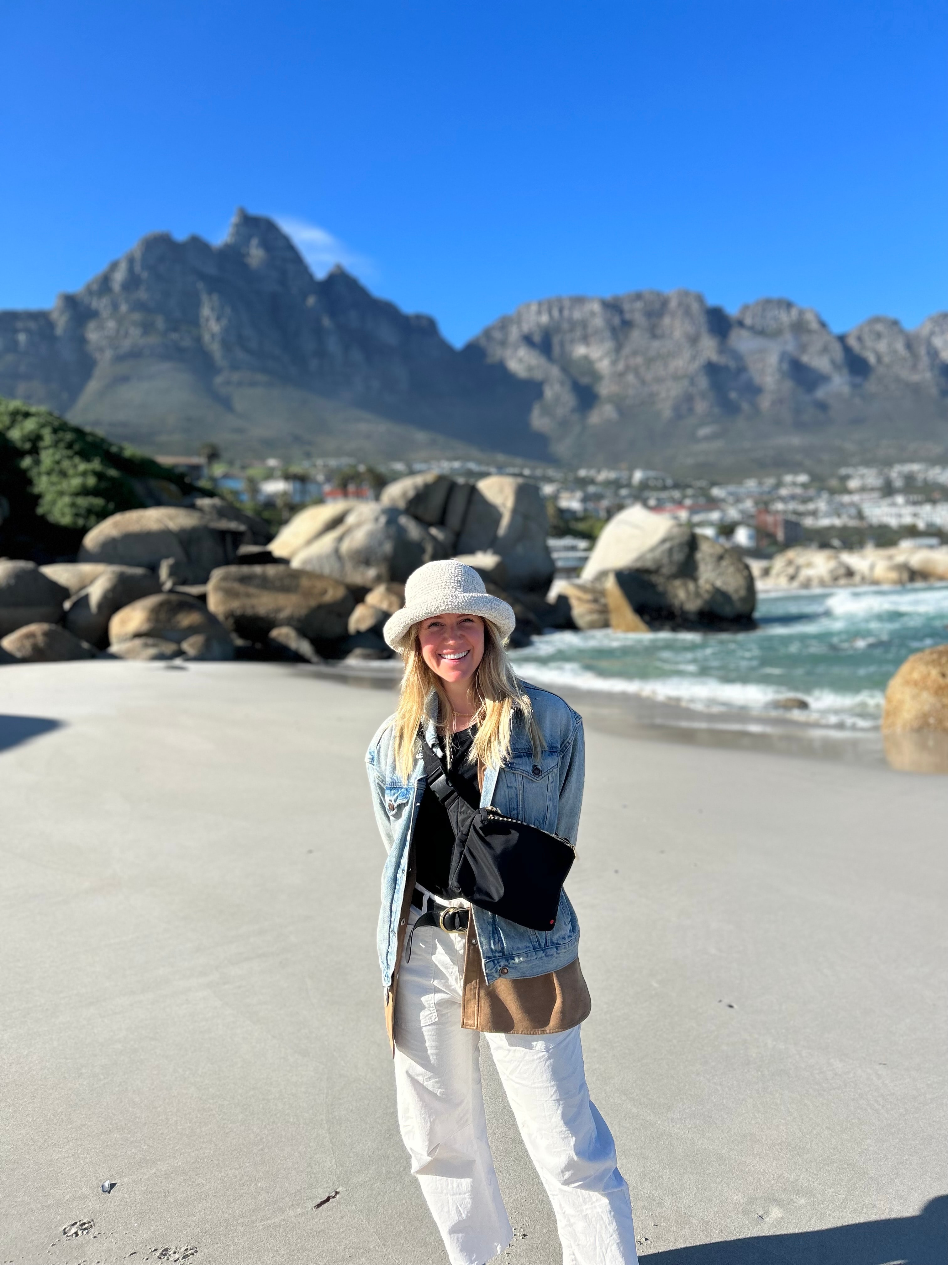 Molly in a white bucket hat standing on a beautiful empty beach with a mountain landscape visible behind her and clear skies