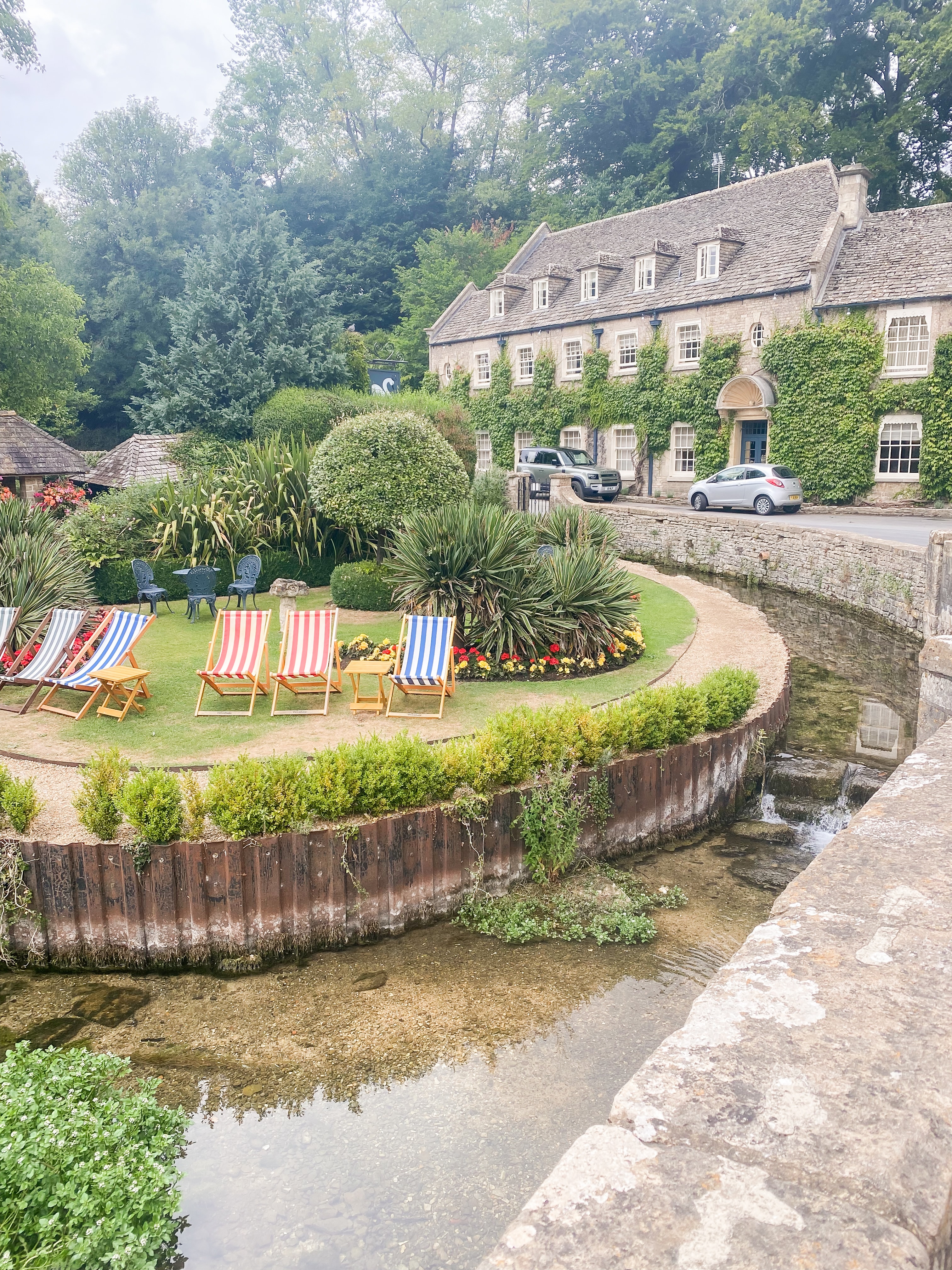 A view of lawn chairs lined up above an empty canal with large brick buildings and a trees in the background during the day.