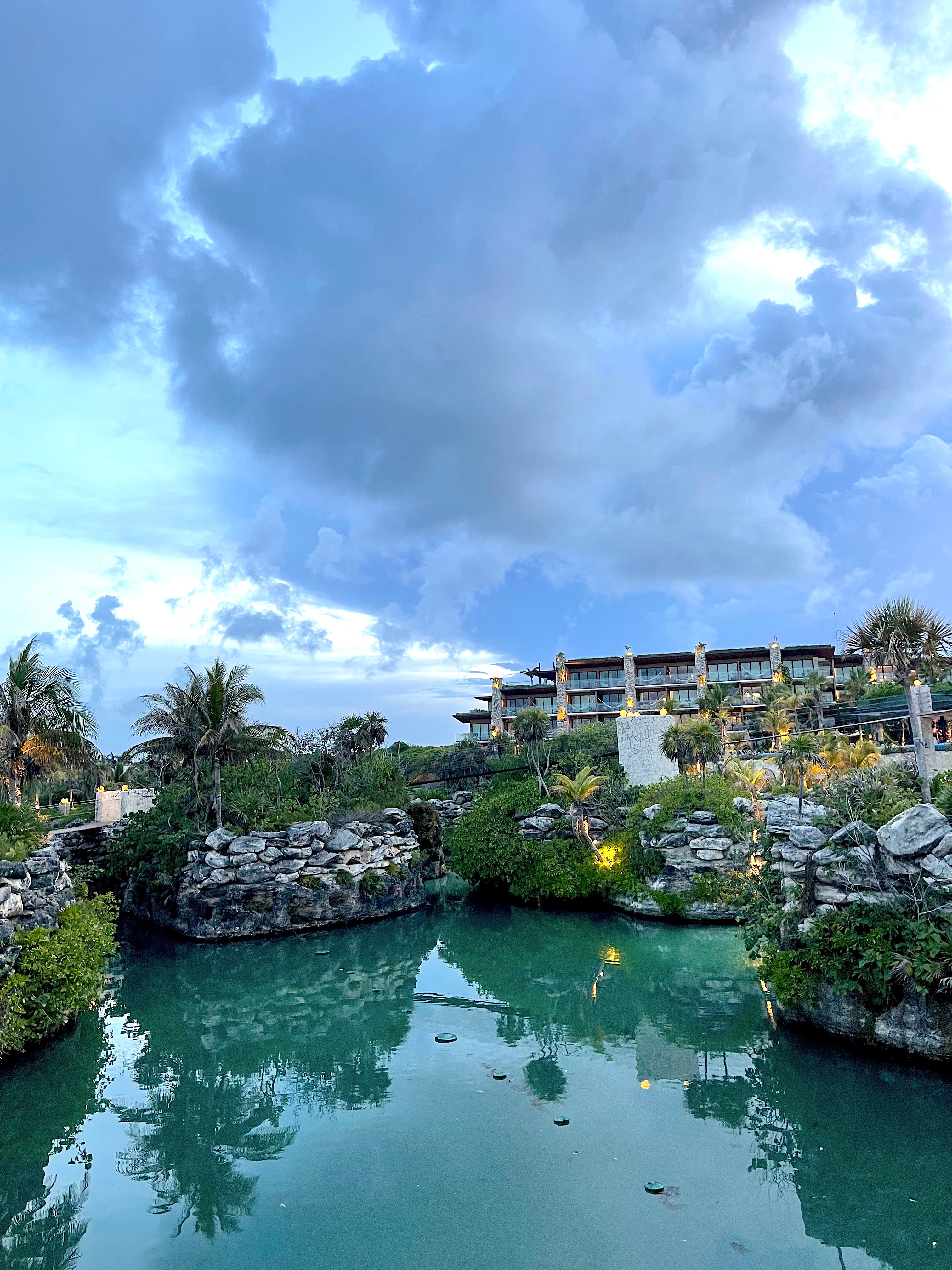 View of a resort pool lined with plants and a building visible in the distance on a cloudy day