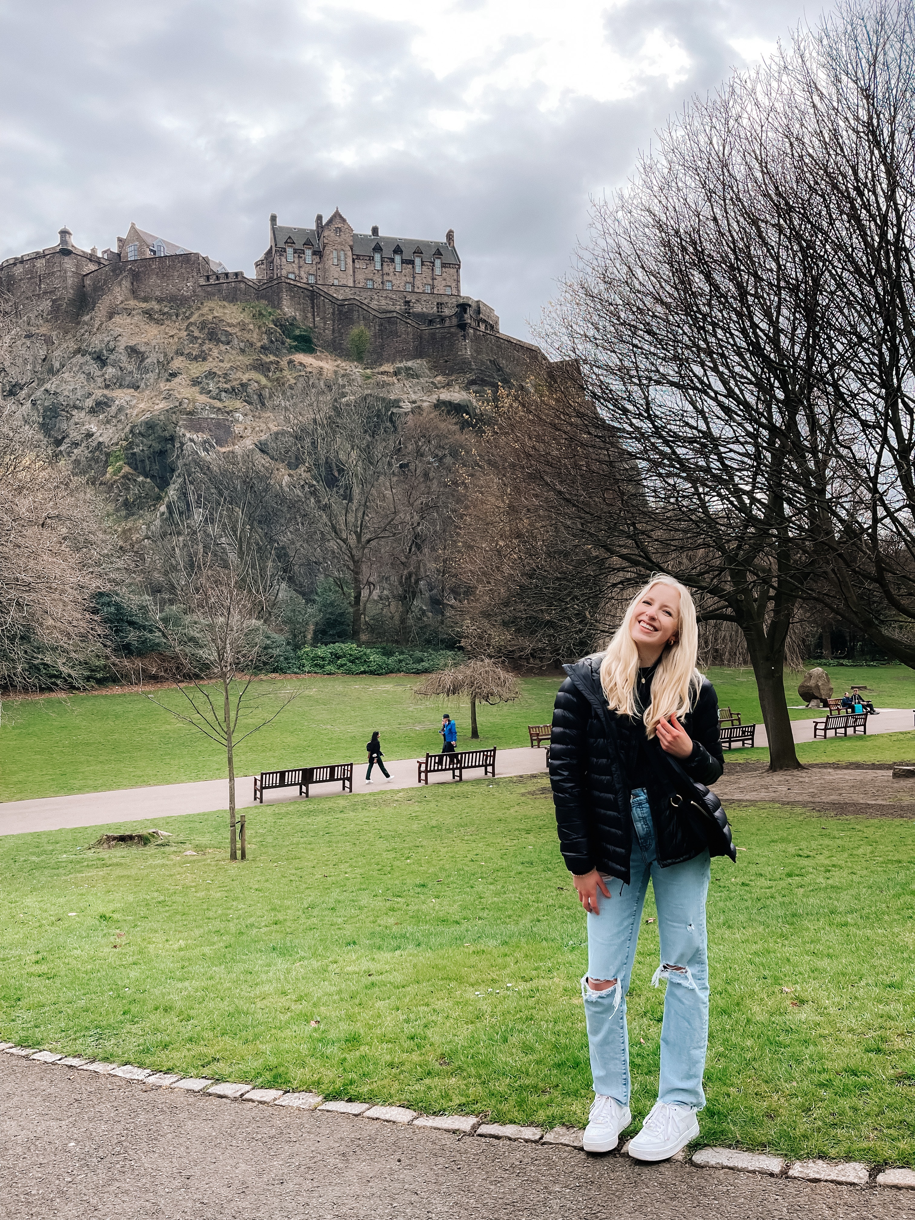 Ellie in a black coat and jeans posing in a park with Edinburgh castle visible behind her