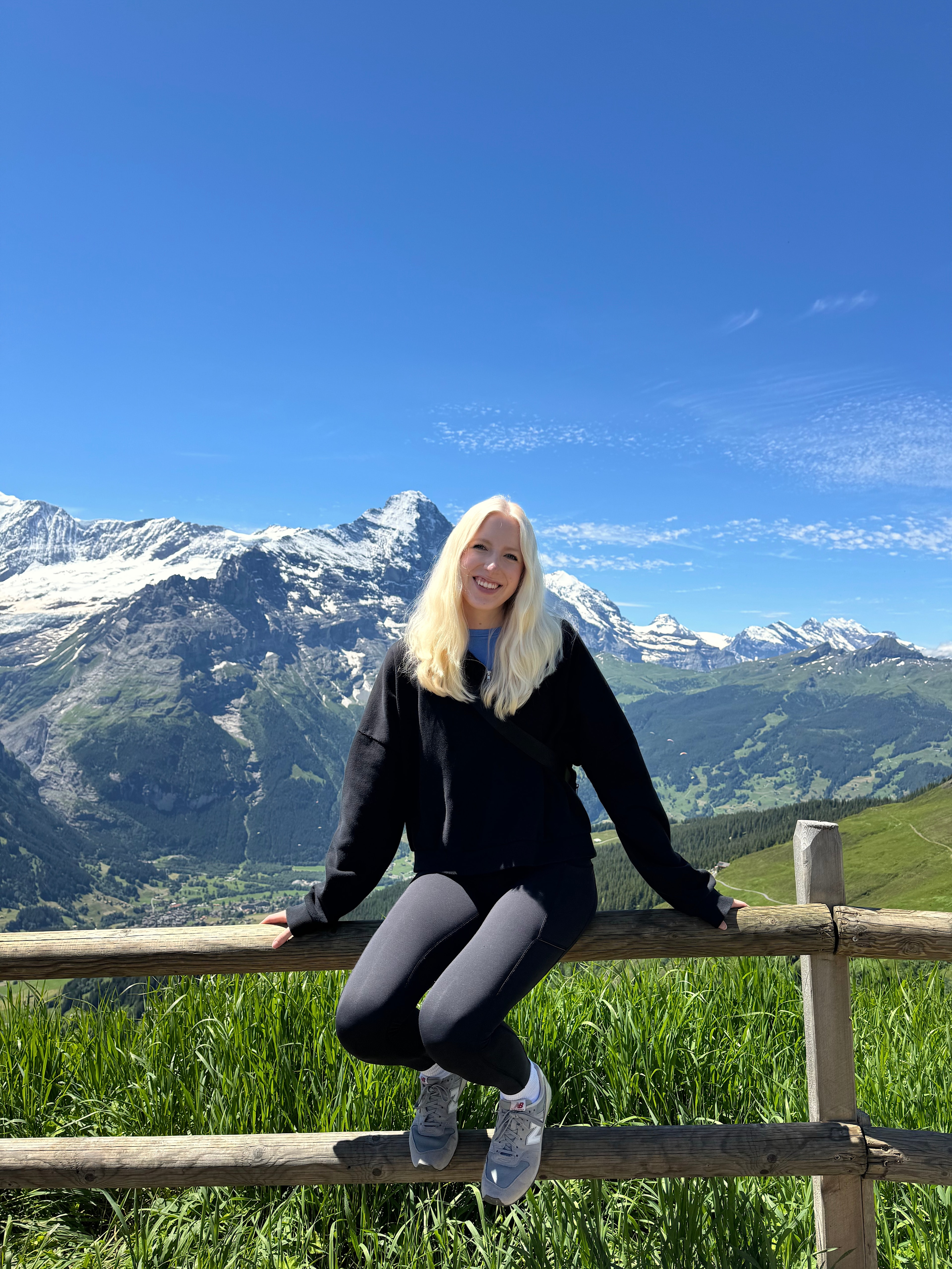 Ellie in all black sitting on a wooden fence overlooking a beautiful snowy mountain landscape on a clear day