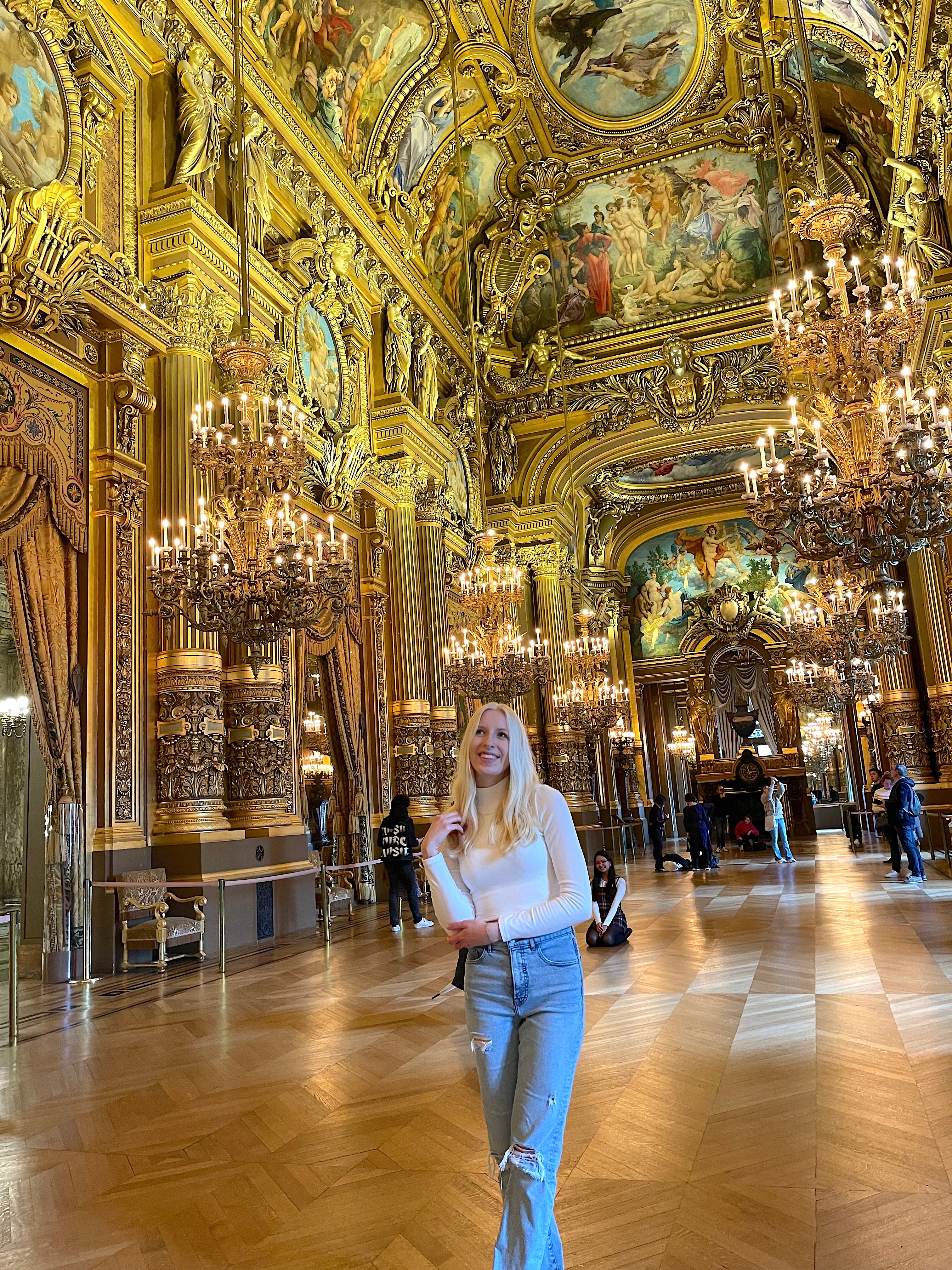 Ellie in an elaborately decorated golden room with large chandeliers