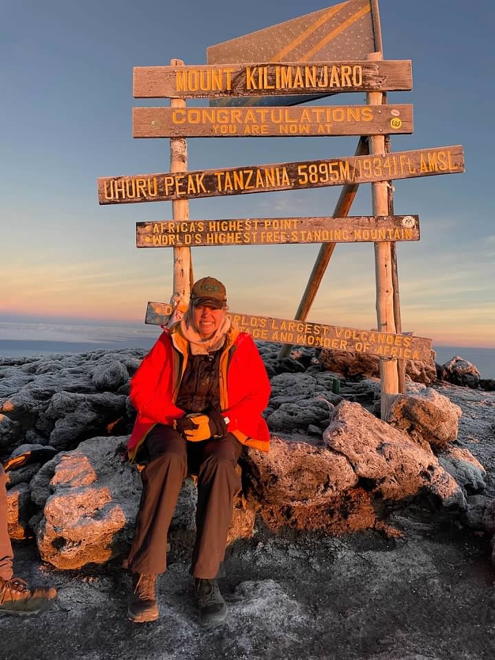 Advisor in a winter jacket sitting on rocks by signposts for Mt. Kilimanjaro under clear skies 