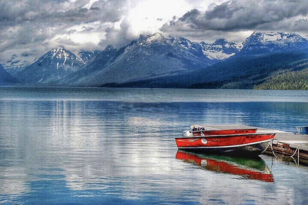 A boat rests on still waters as clouds cascade down a mountainside on the shore. 