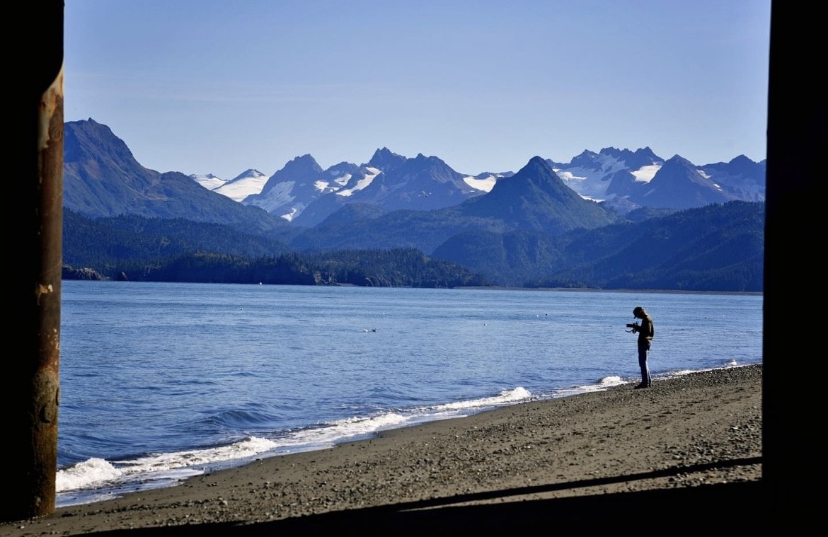 A mountain range stands on the horizon as waves gently lap the shore on a clear day. 