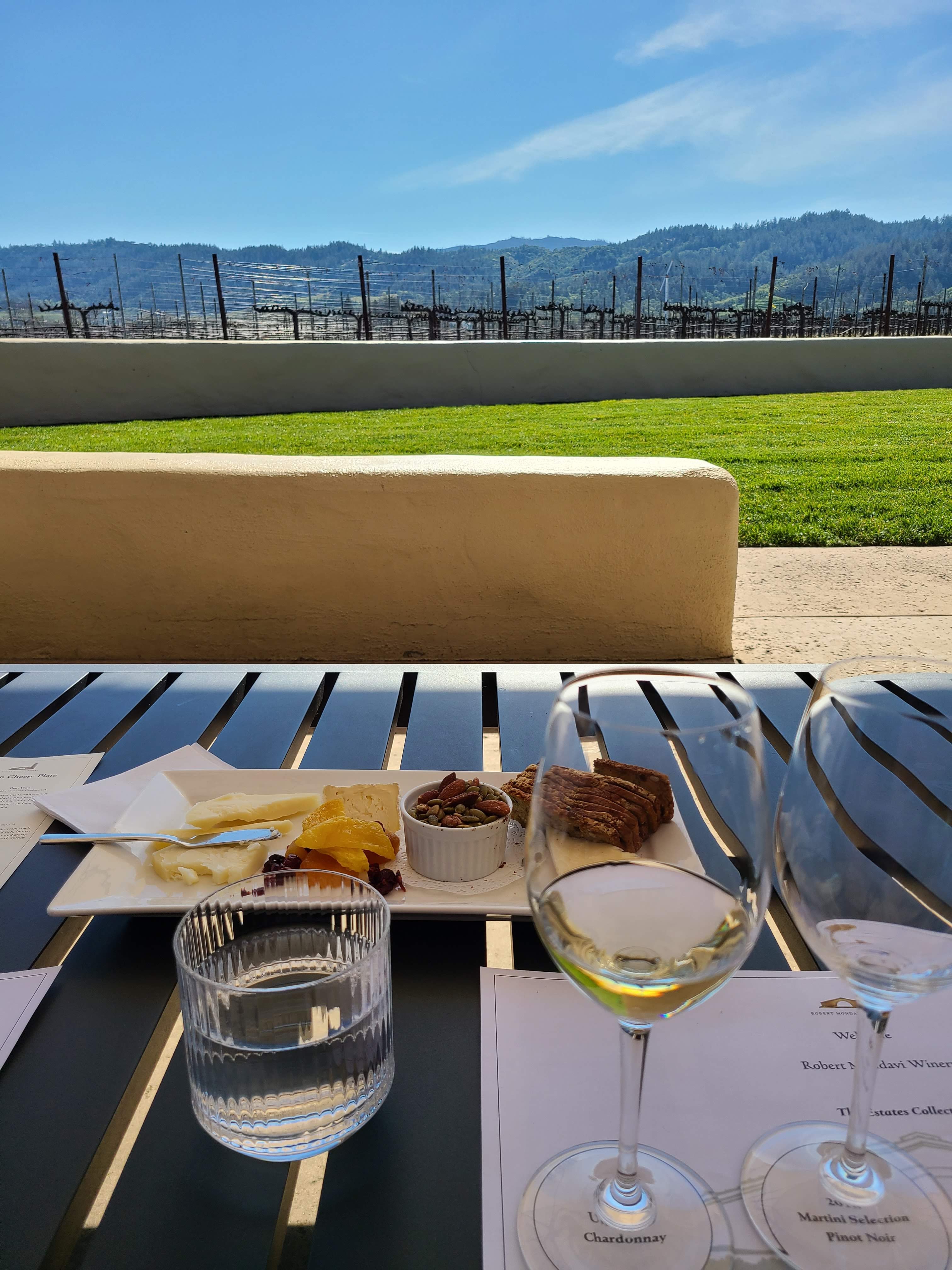 View of two glasses of wine and a cheese plate set on an outdoor table at a winery on a sunny day