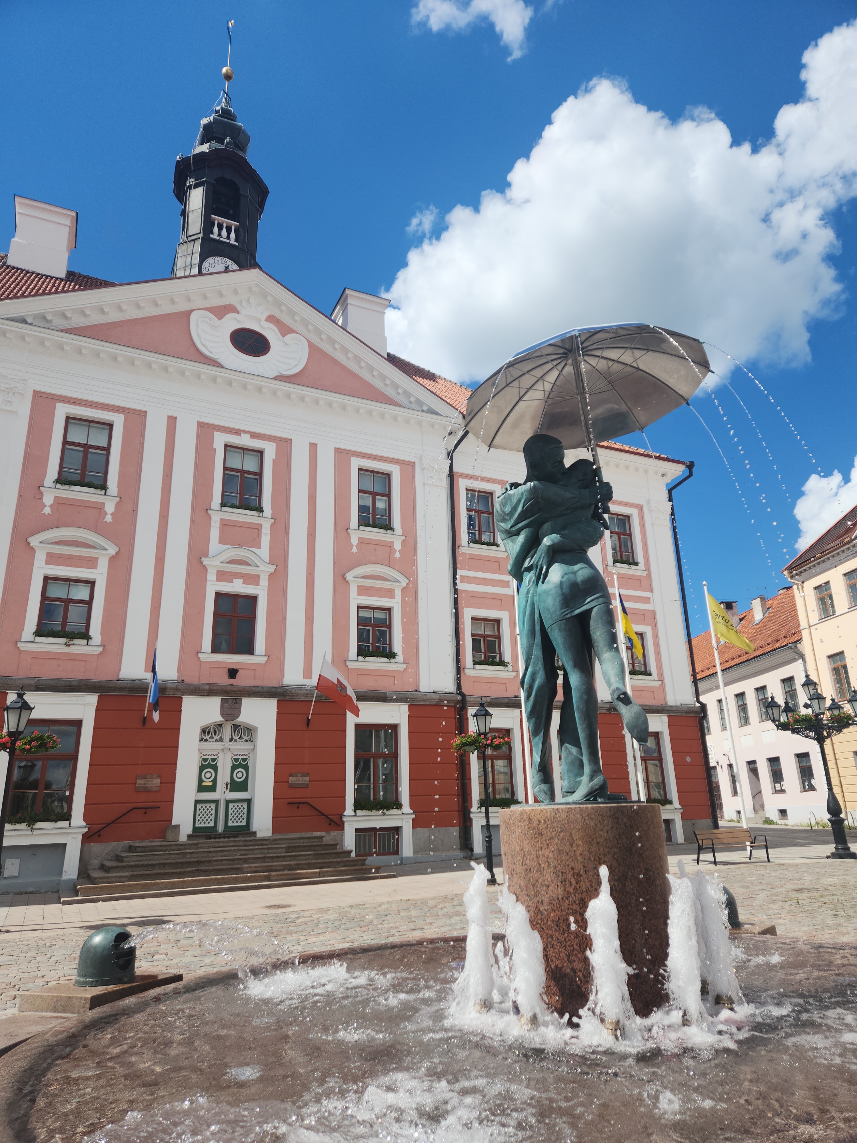 View of a statue fountain of two people embracing in front of a pink building on a sunny day