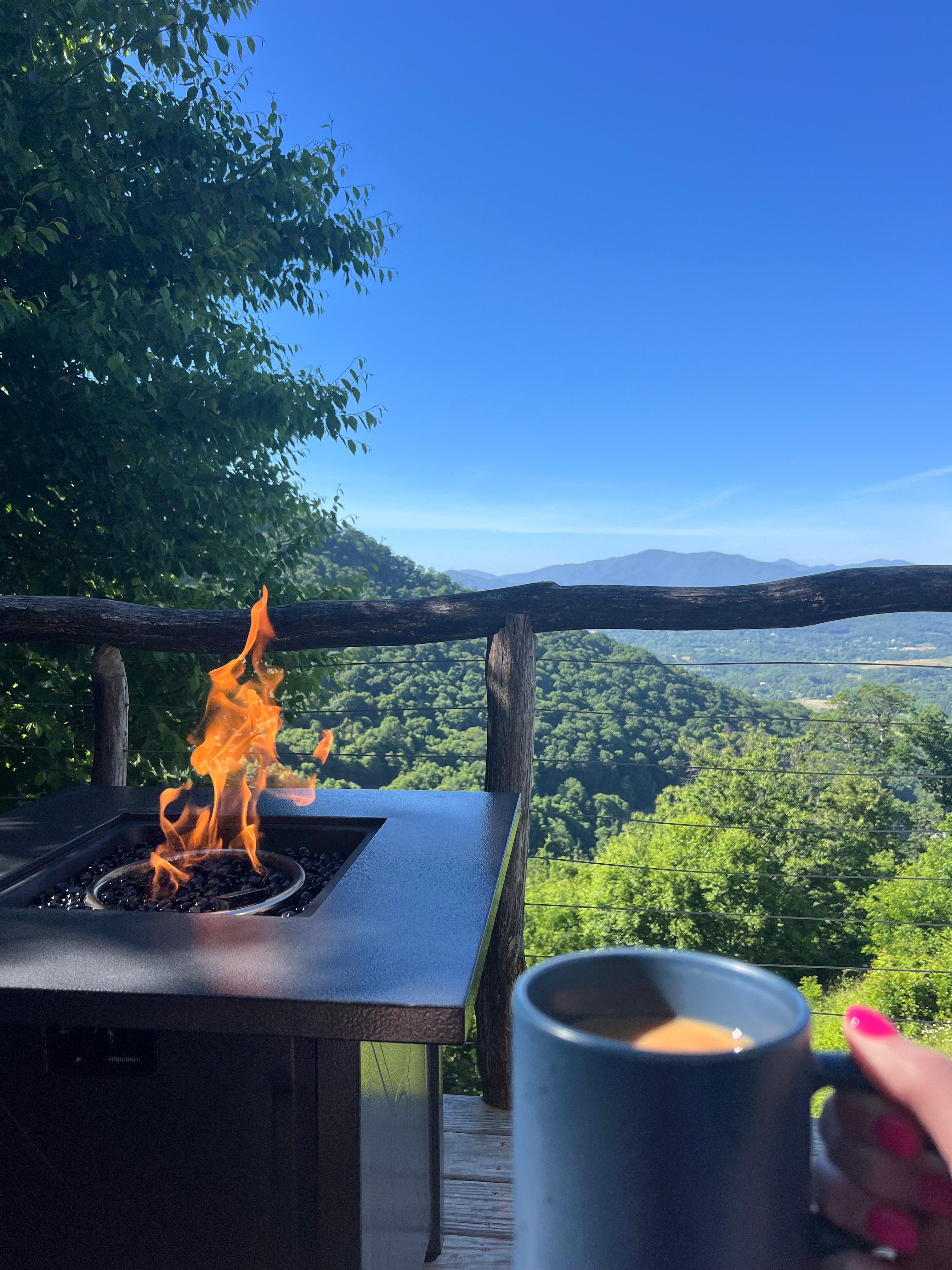 View of a hand holding a mug of coffee outdoors on a terrace with a fire pit overlooking a green valley and blue skies