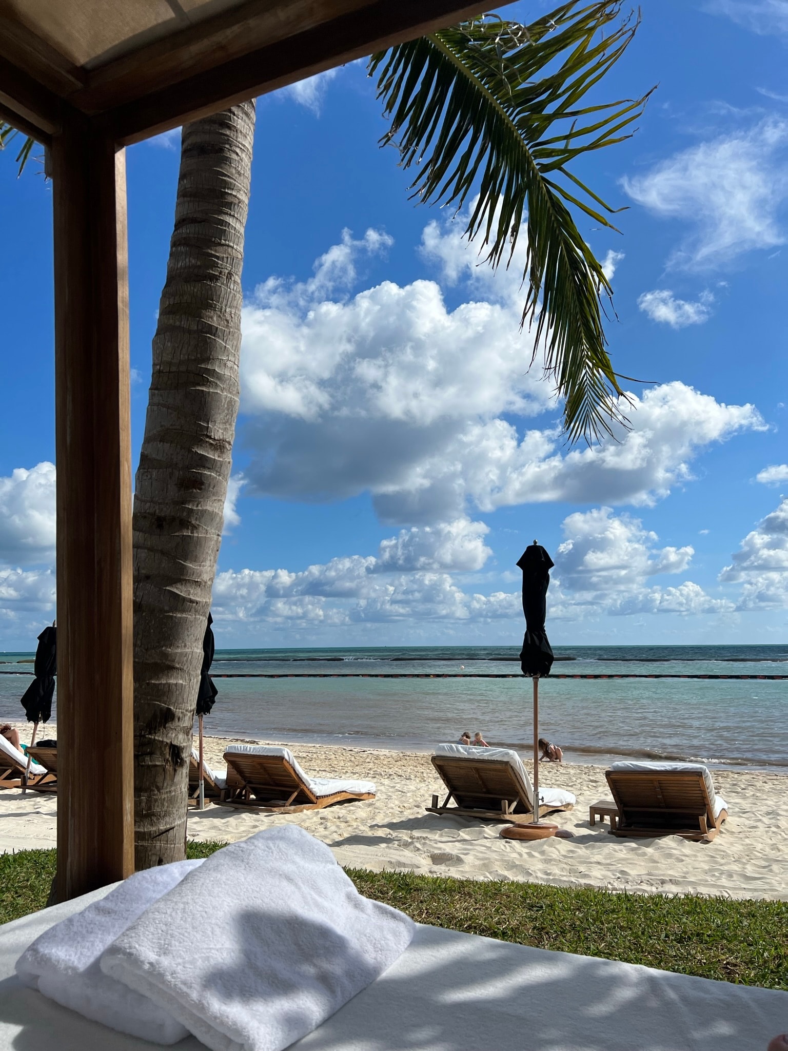 A view from a cabana of a white sand beach with lounge chairs, palm trees and a calm ocean in the distance.