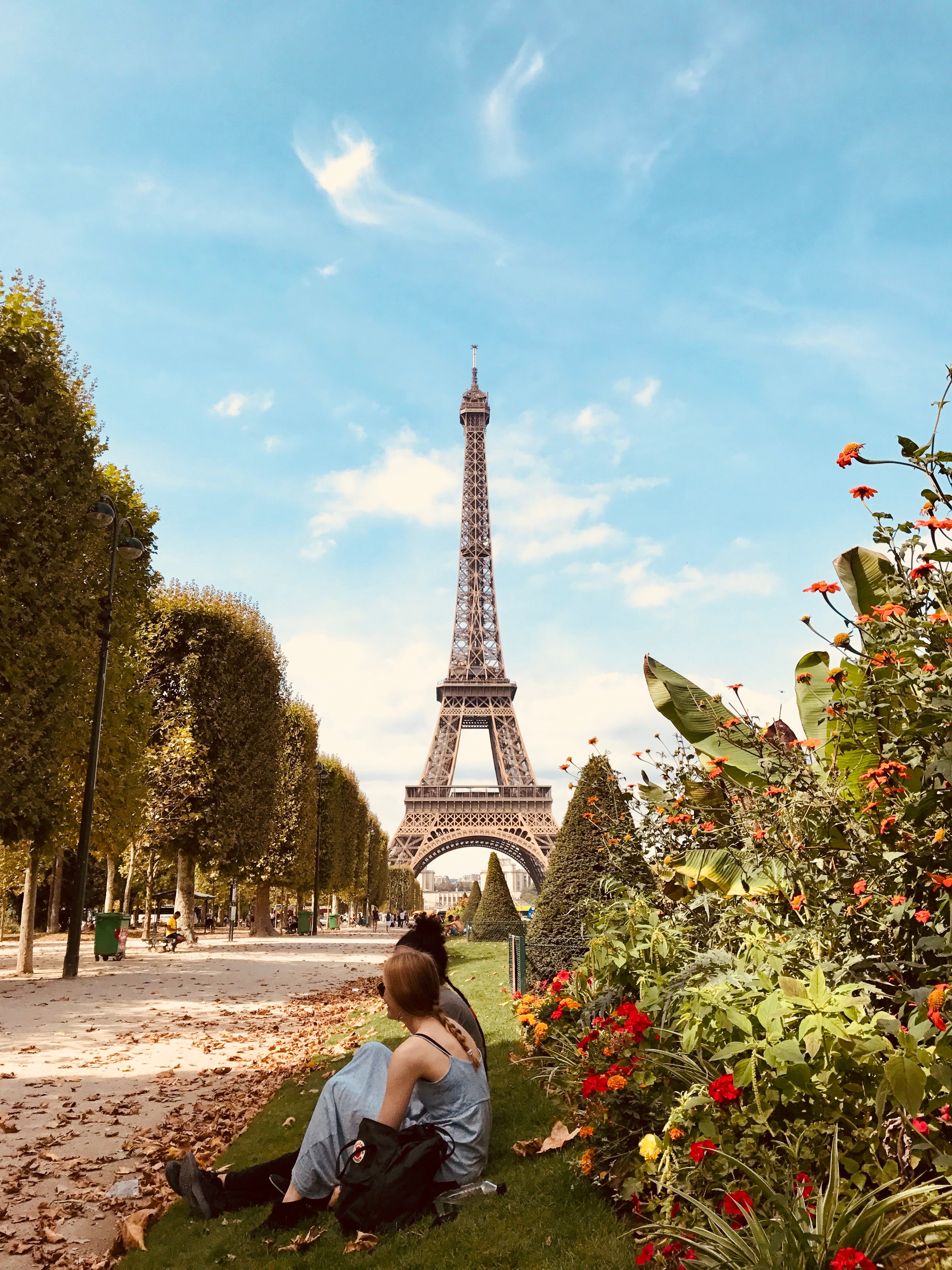 A couple poses on a grassy patch surrounded by flowers as the Eiffel Tower reaches for a clear blue sky dotted with clouds. 