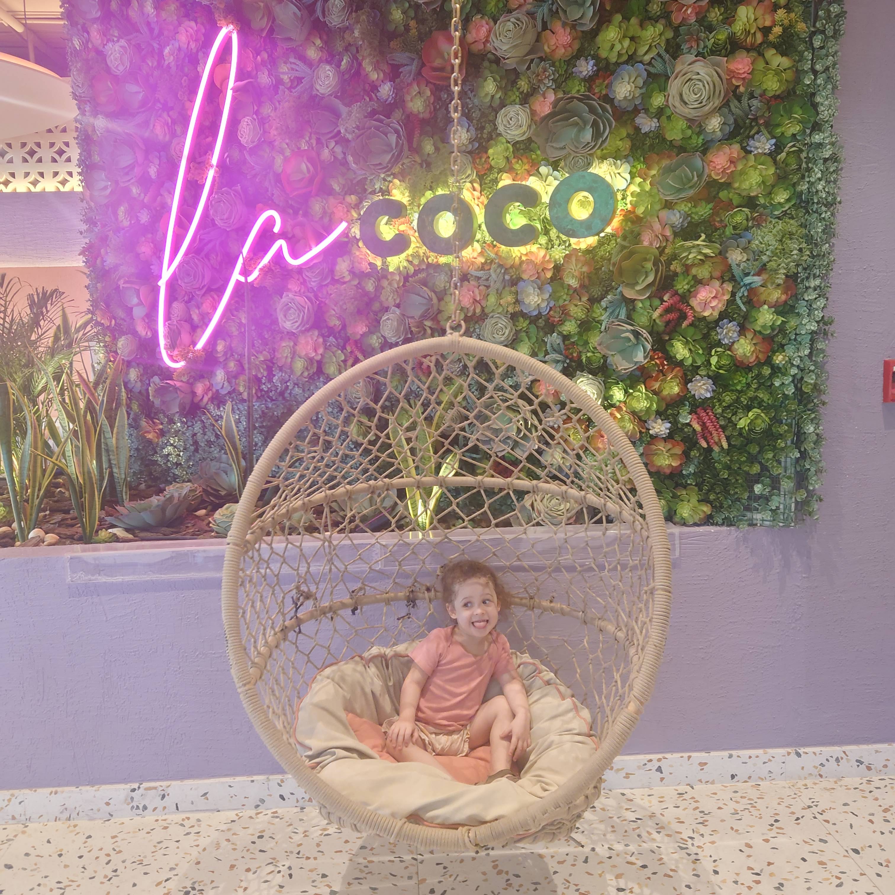 Young girl sitting in a round swing chair in front of a neon sign reading “la coco”