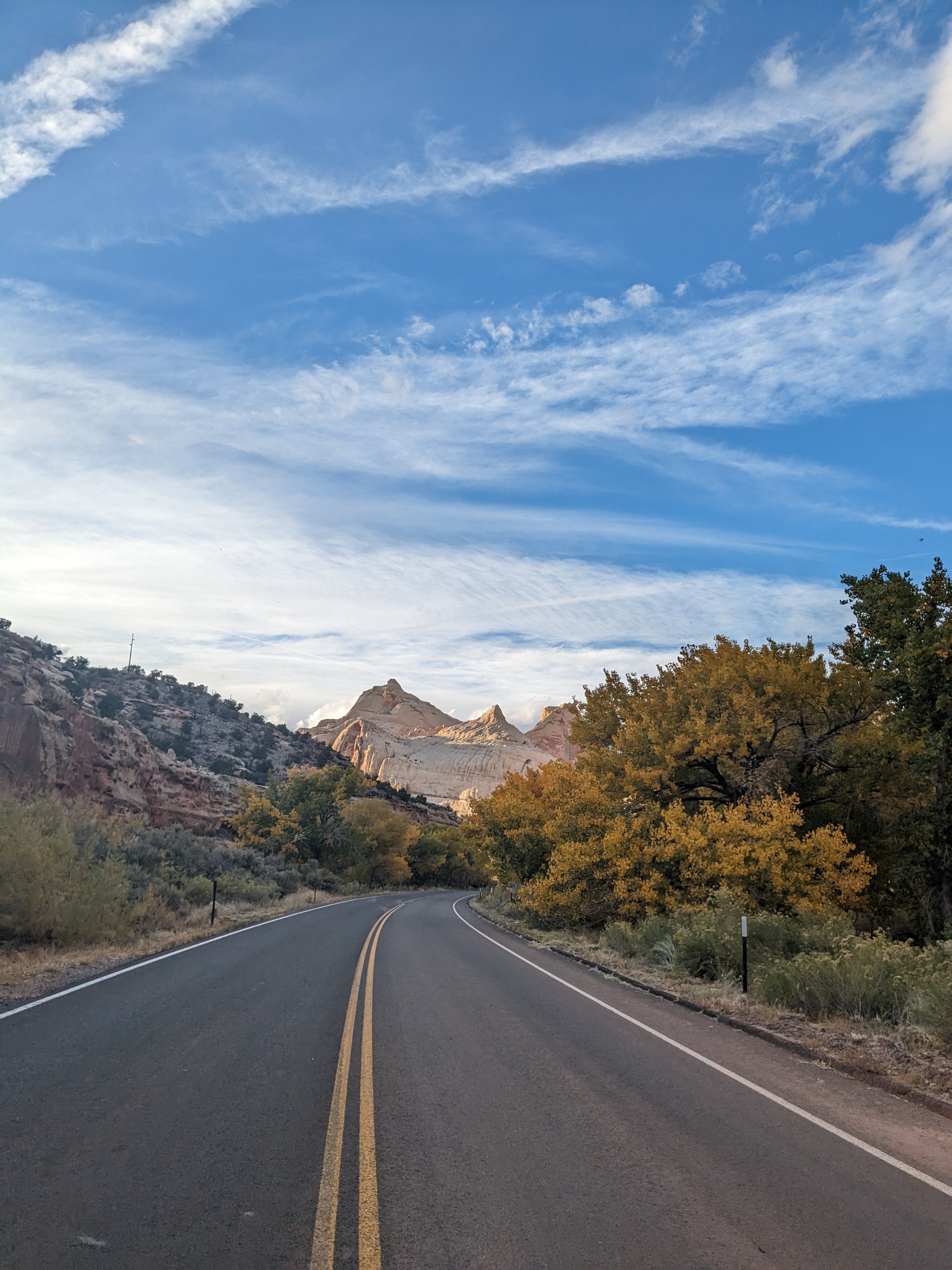 An open road with mountains and natural foliage in surrounding view beneath a blue sky with scattered clouds. 