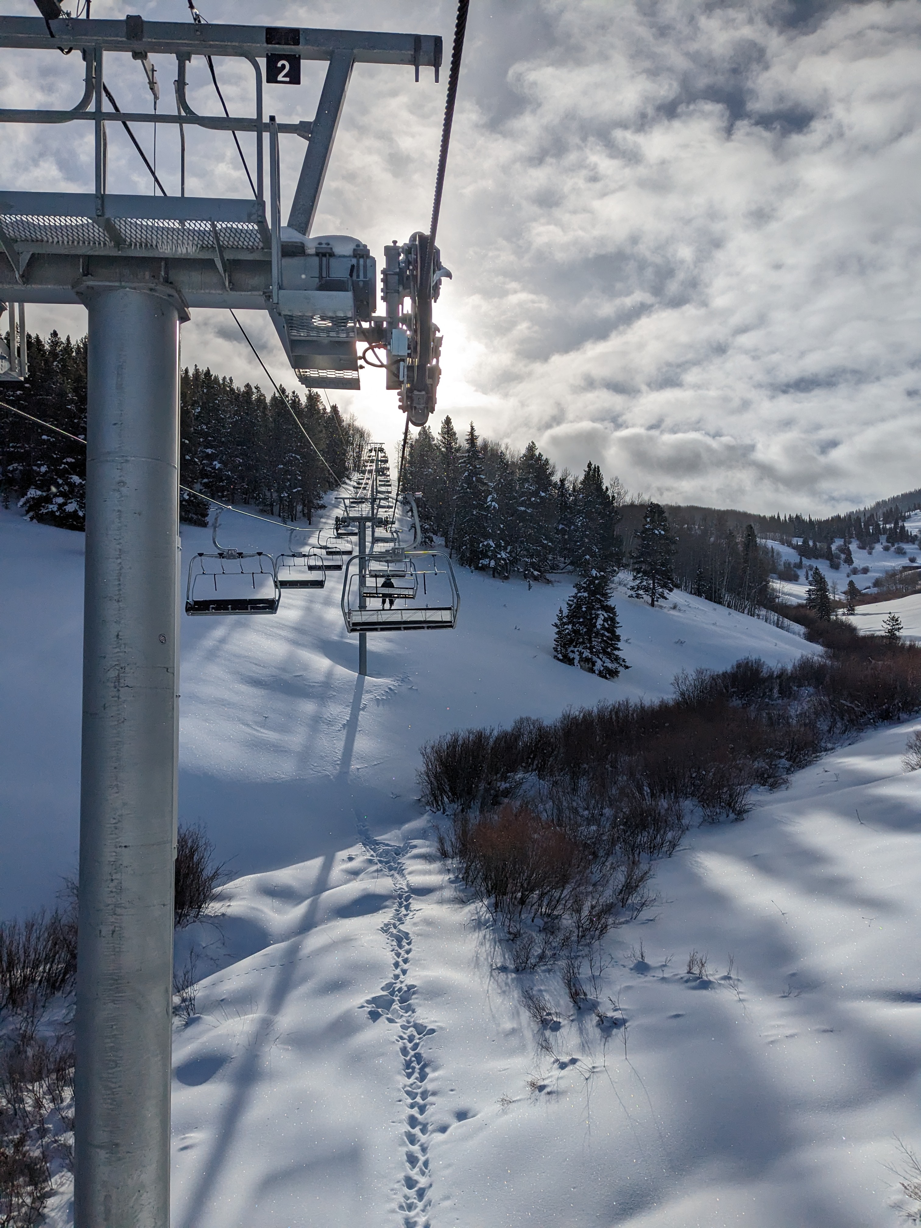 A ski lift towering above snowy hills and pine trees with scattered clouds and a hint of the sun shining above it. 