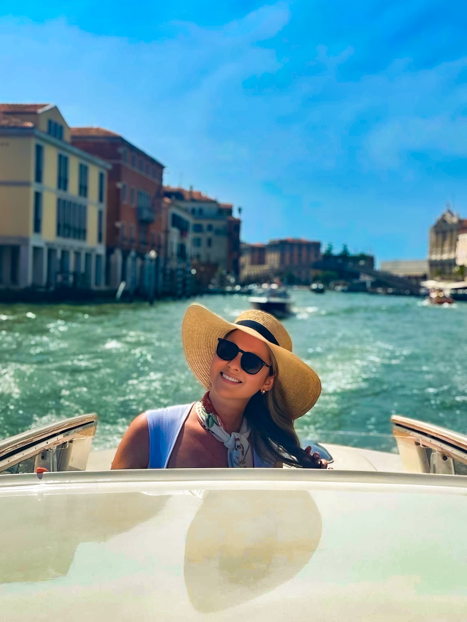 A woman wearing a hat and sunglasses enjoys a sunny day on a boat, surrounded by calm water.