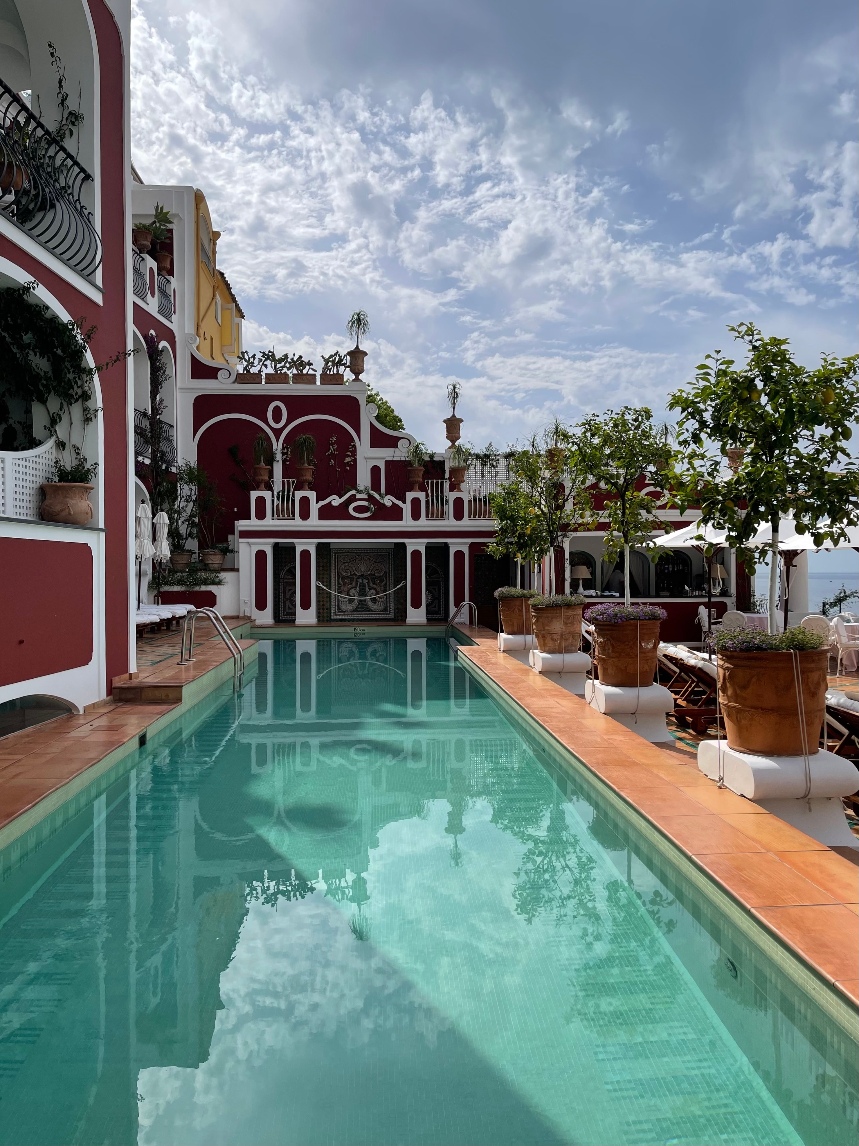 View of a long empty hotel swimming pool next to a red building with white trim on a sunny day