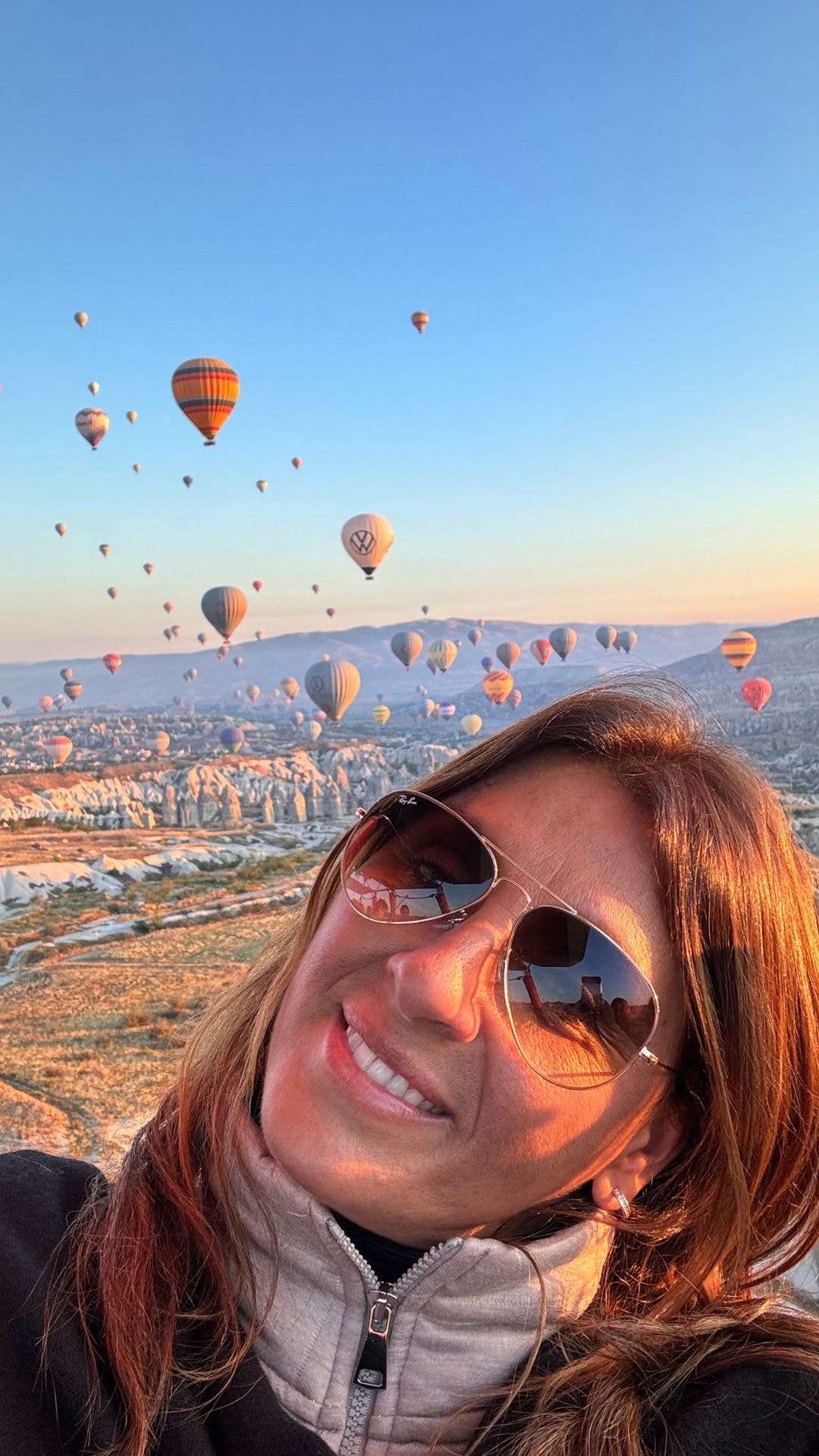 Advisor in sunglasses taking a selfie with the many hot air balloons of Cappadocia, Turkey in flight behind her