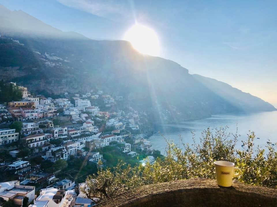 View of the sun shining over mountains on the coast with a small coastal town on the hill below