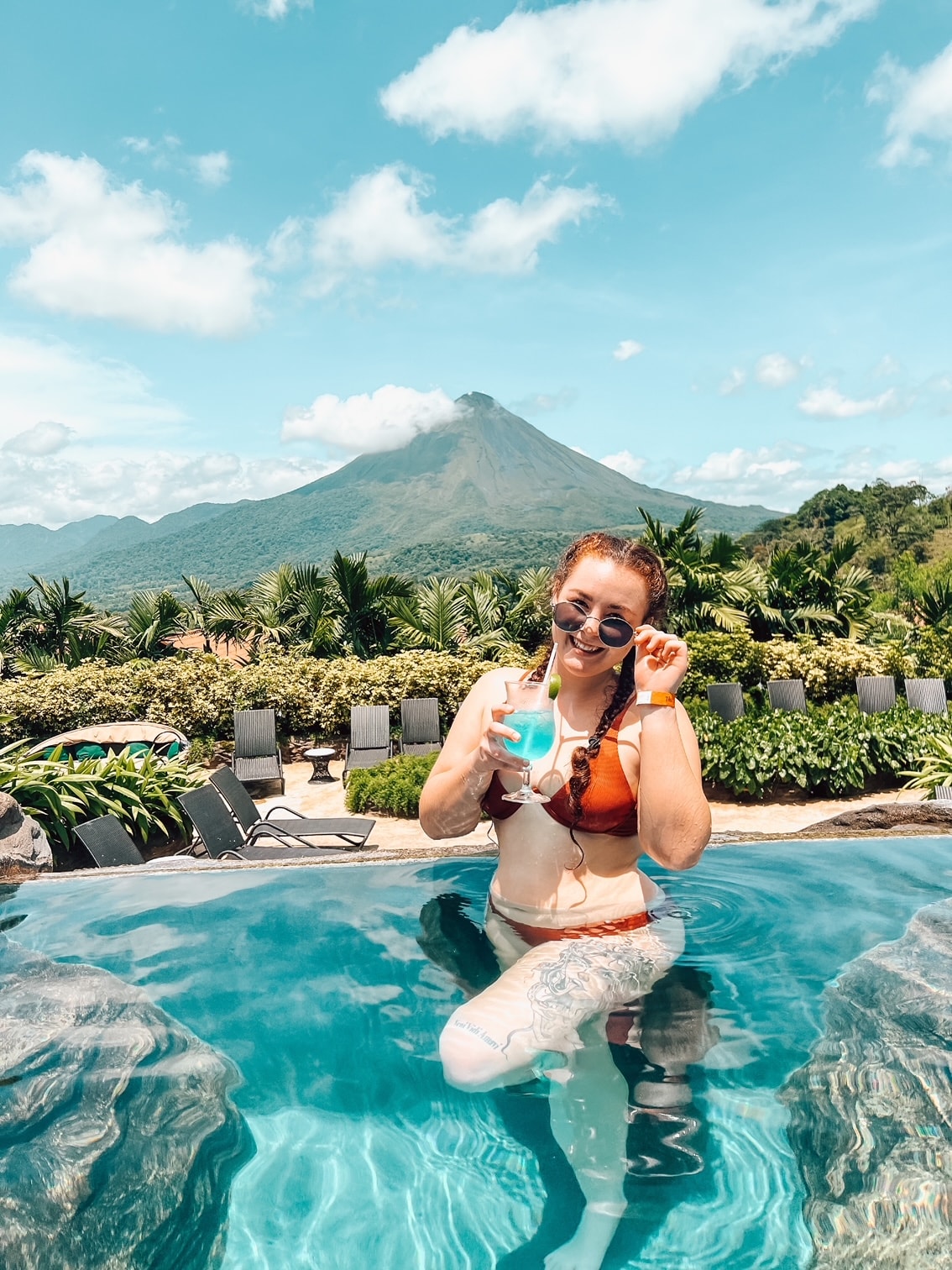 Advisor posing in an infinity pool with blue cocktail in hand overlooking a lush landscape and a volcano rising in the distance