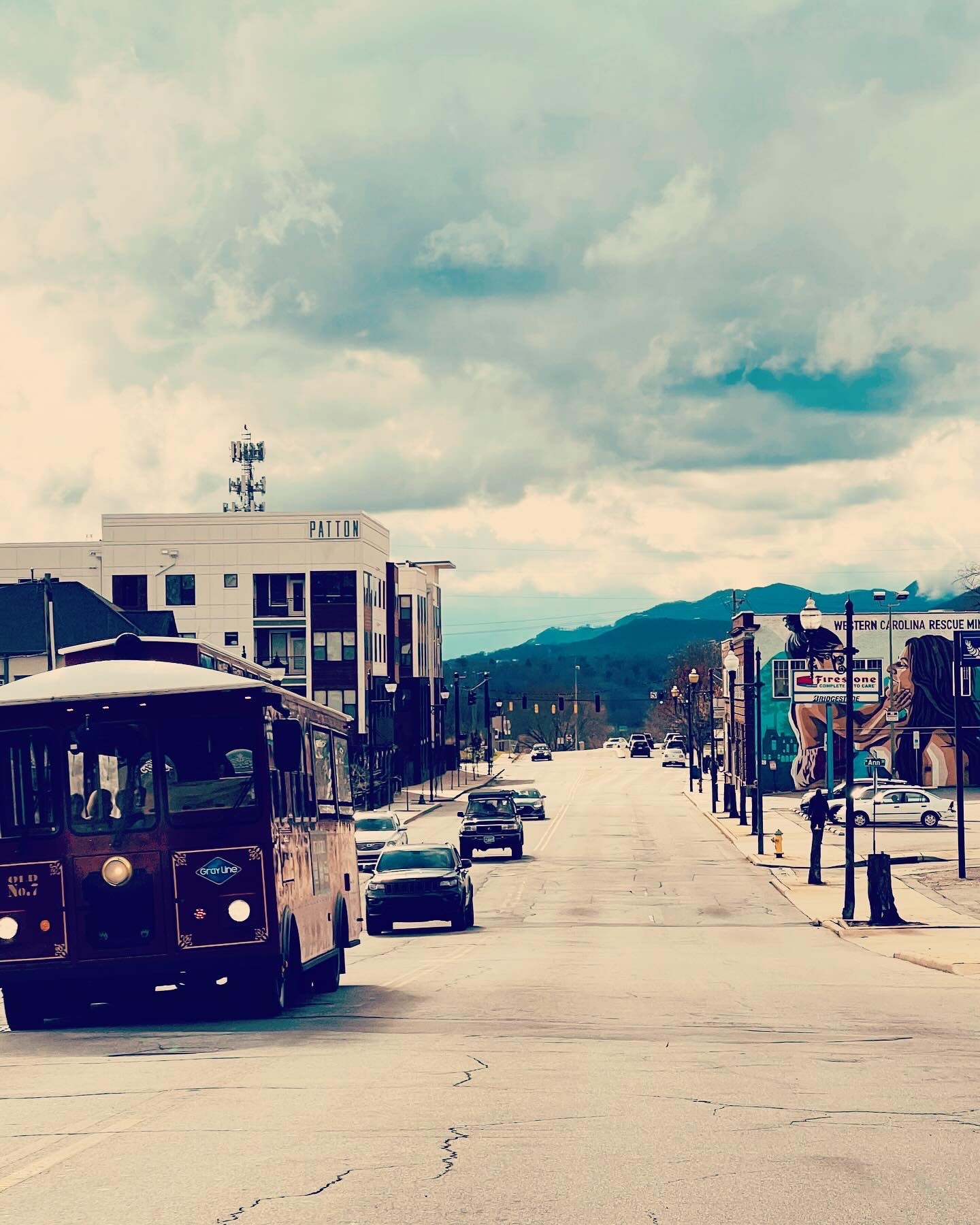 View of a small-town road with cars driving towards the camera on a cloudy day with mountains visible in the distance