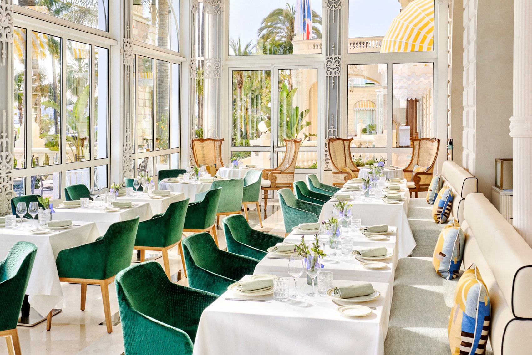 a luxe sunny restaurant with green velvet chairs around tables draped with white tablecloths.