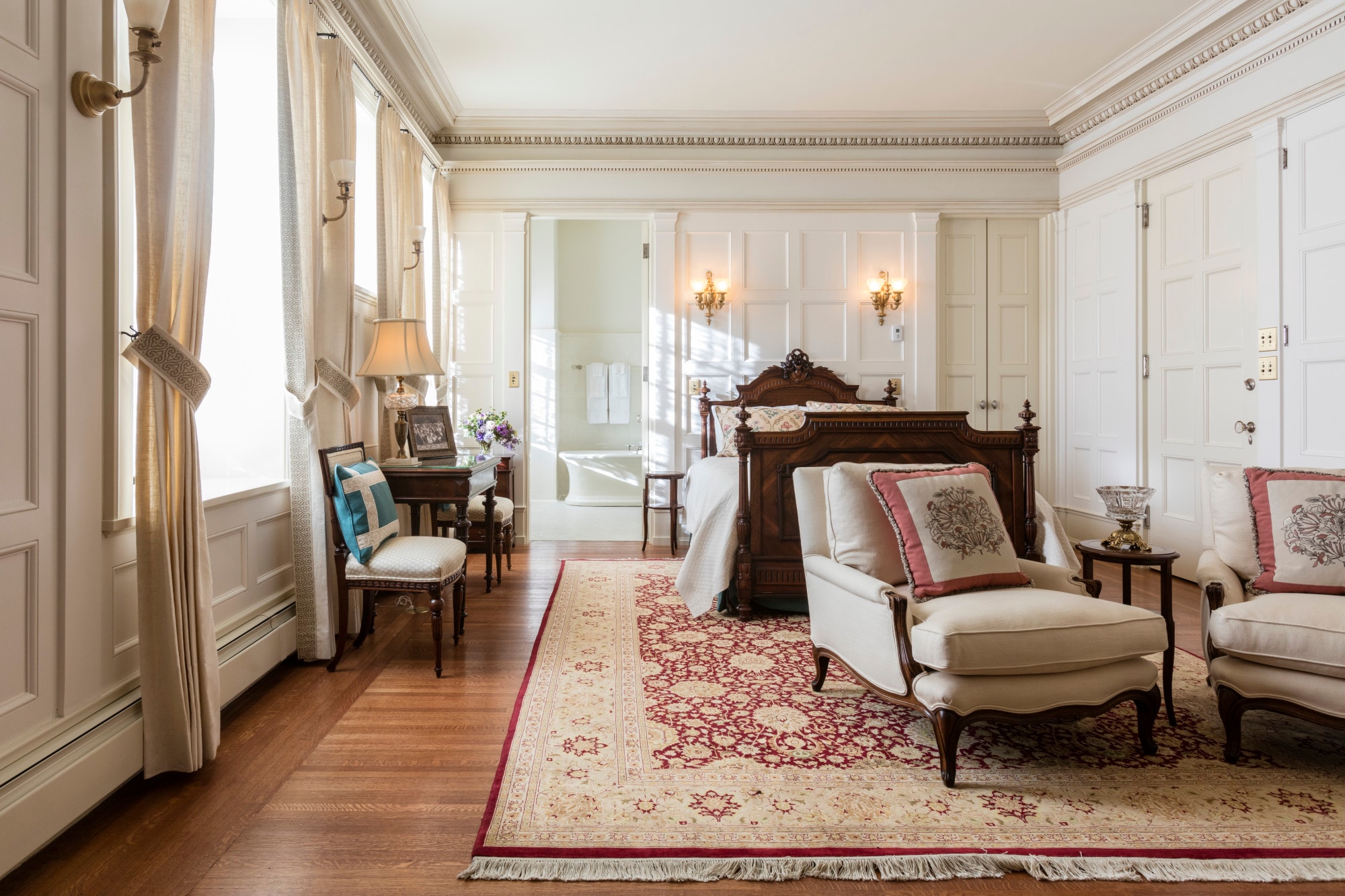 a luxe bedroom with wooden floors covered by an oversized rug, white walls, and crown moulding