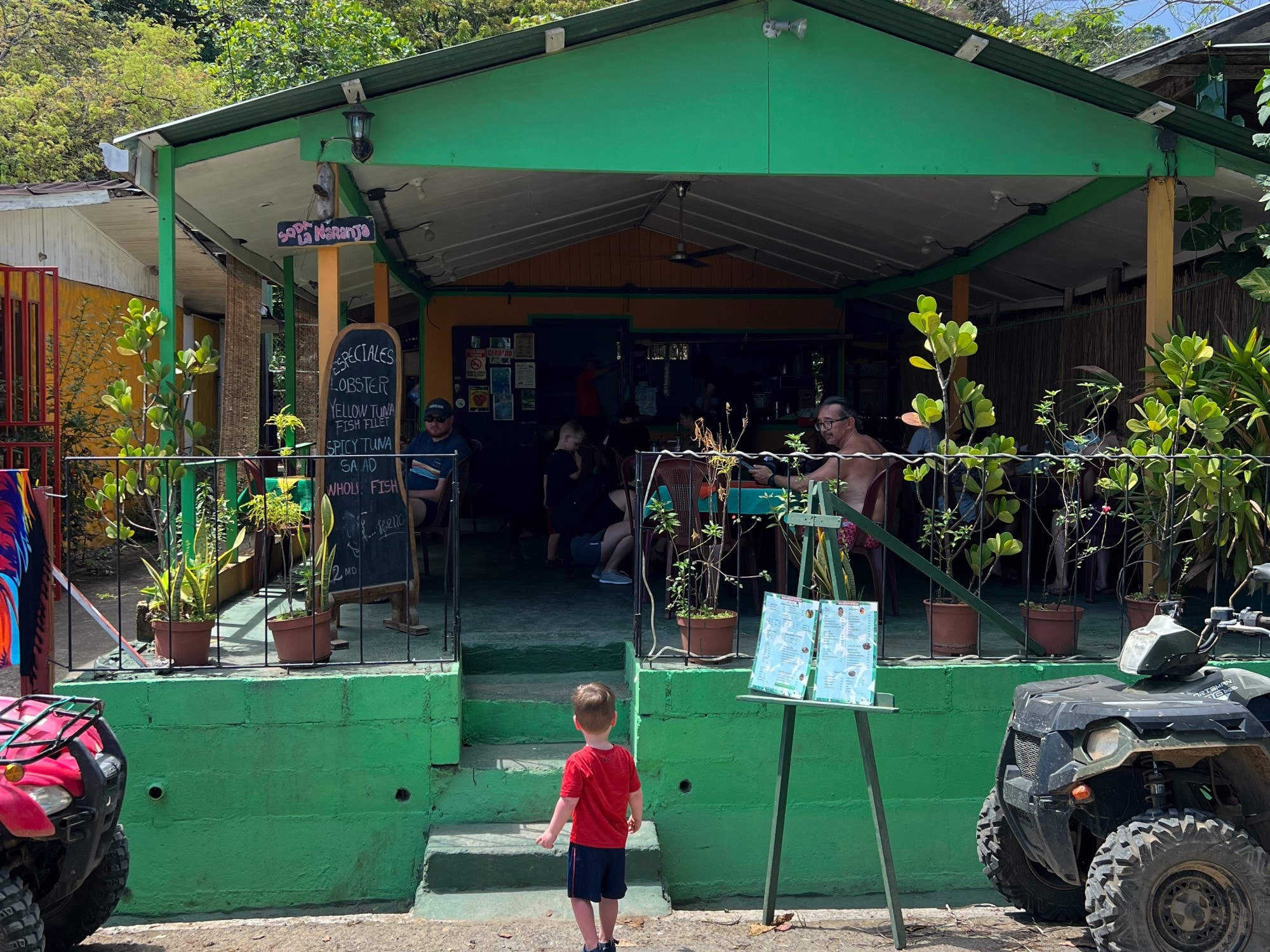 A traditional local restaurant or 'soda' with a green facade and menu board with chalked text.