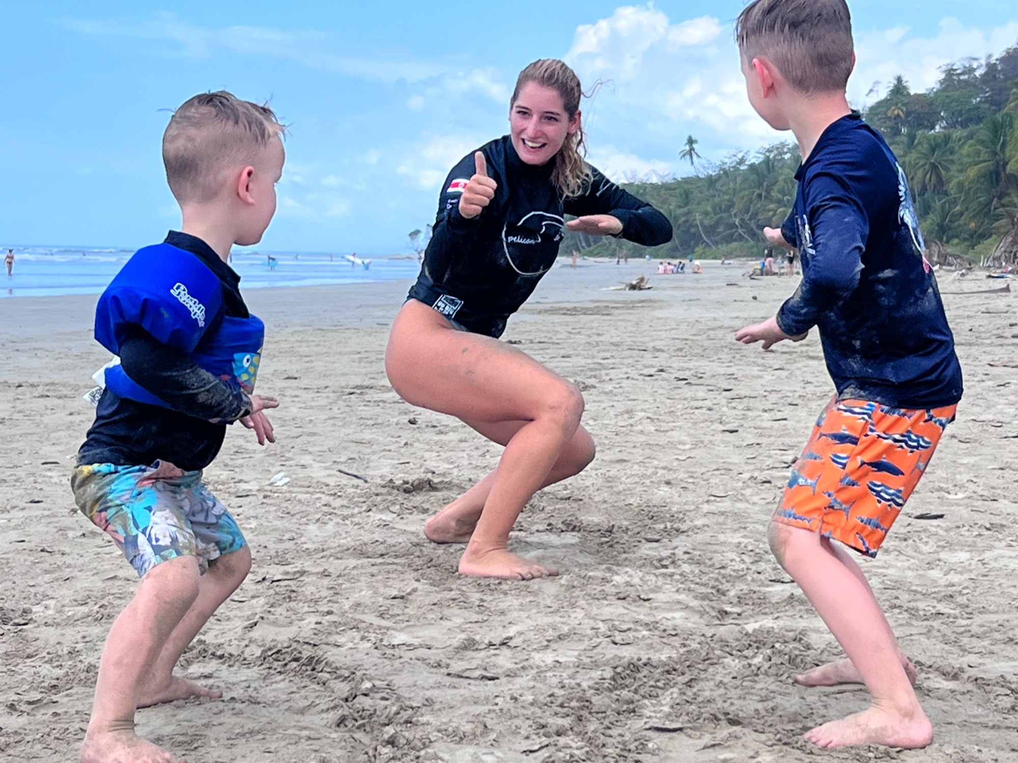 Two children and a woman standing in surfing poses on the beach.