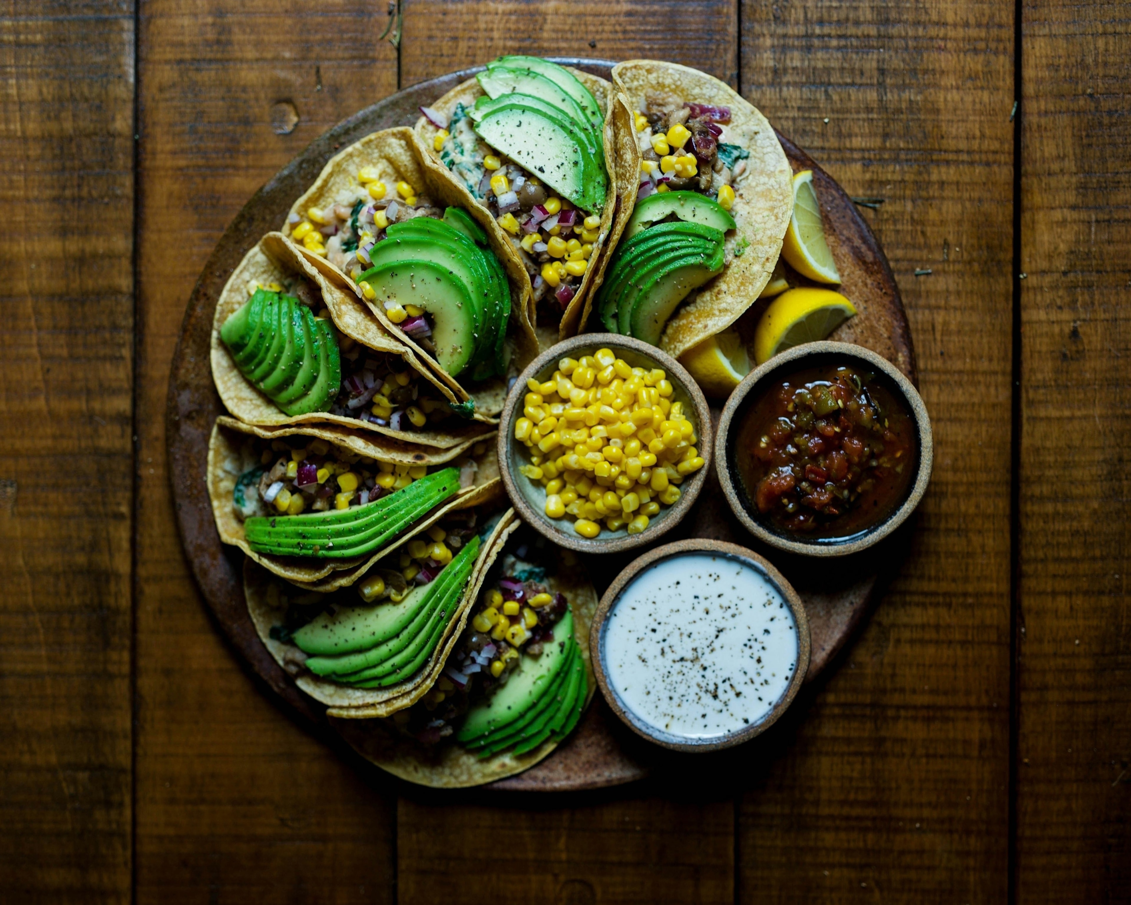 The image shows a wooden table with a plate of tacos filled with avocado slices, corn, and beans, accompanied by two small bowls of salsa and cream.