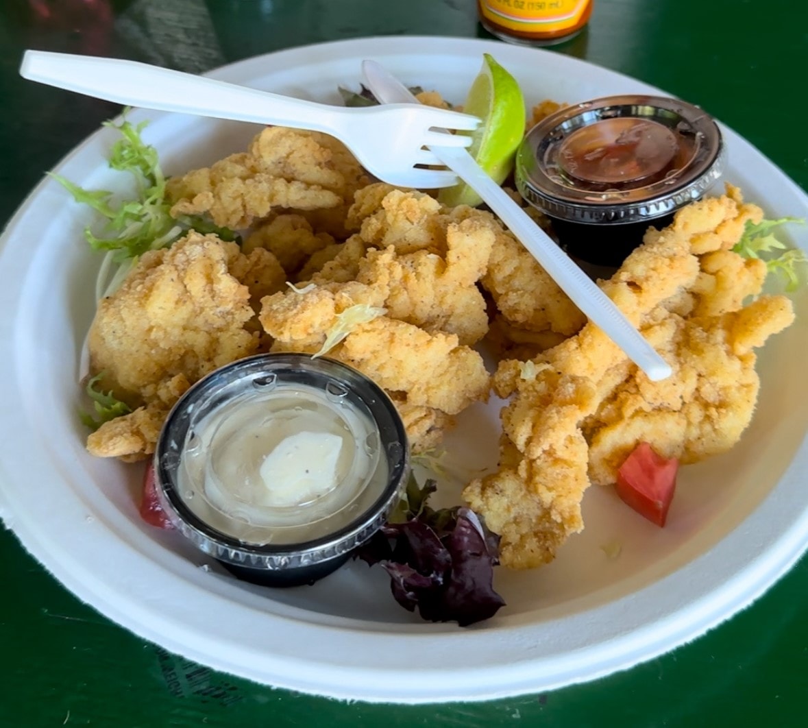 The image features a white plastic plate of food with fried delicacies, with a spicy Mexican salsa bottle to one side.