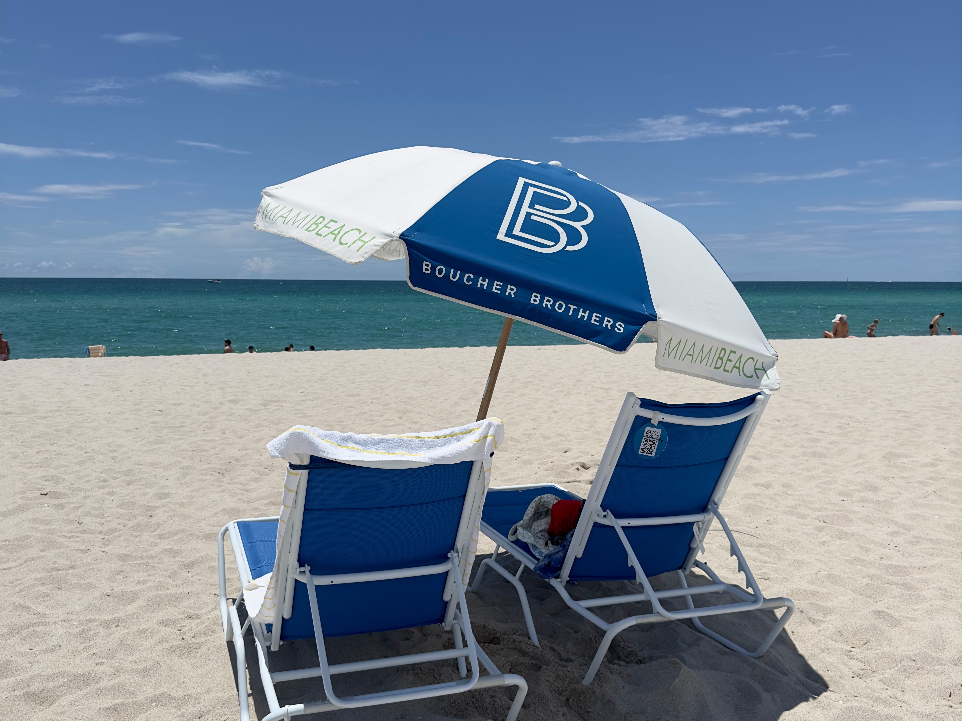 The image features two beach chairs and a “Boucher Brothers” umbrella on a sandy shore.