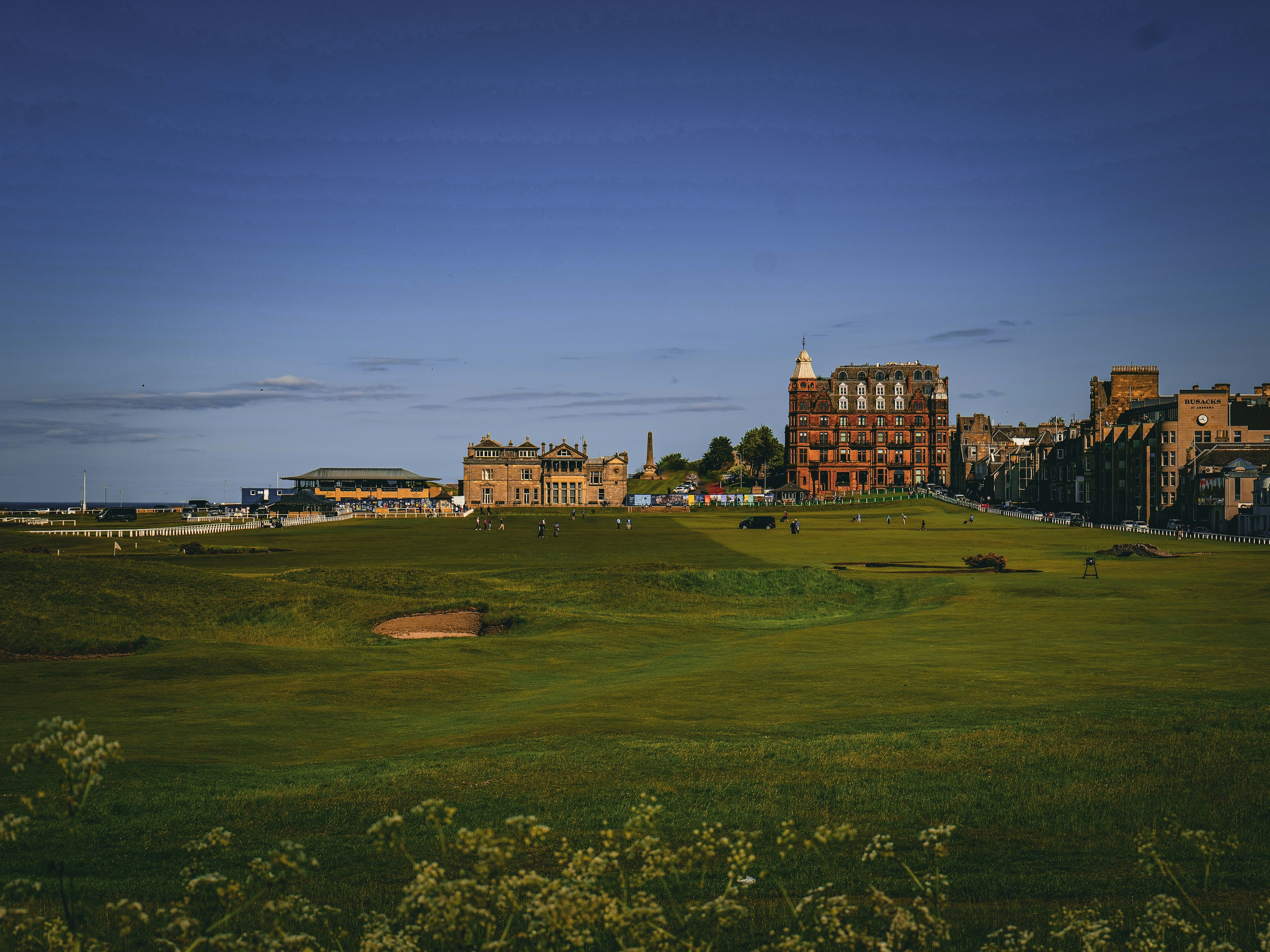 The image shows a picturesque green golf course with buildings in the distance under a clear blue sky.