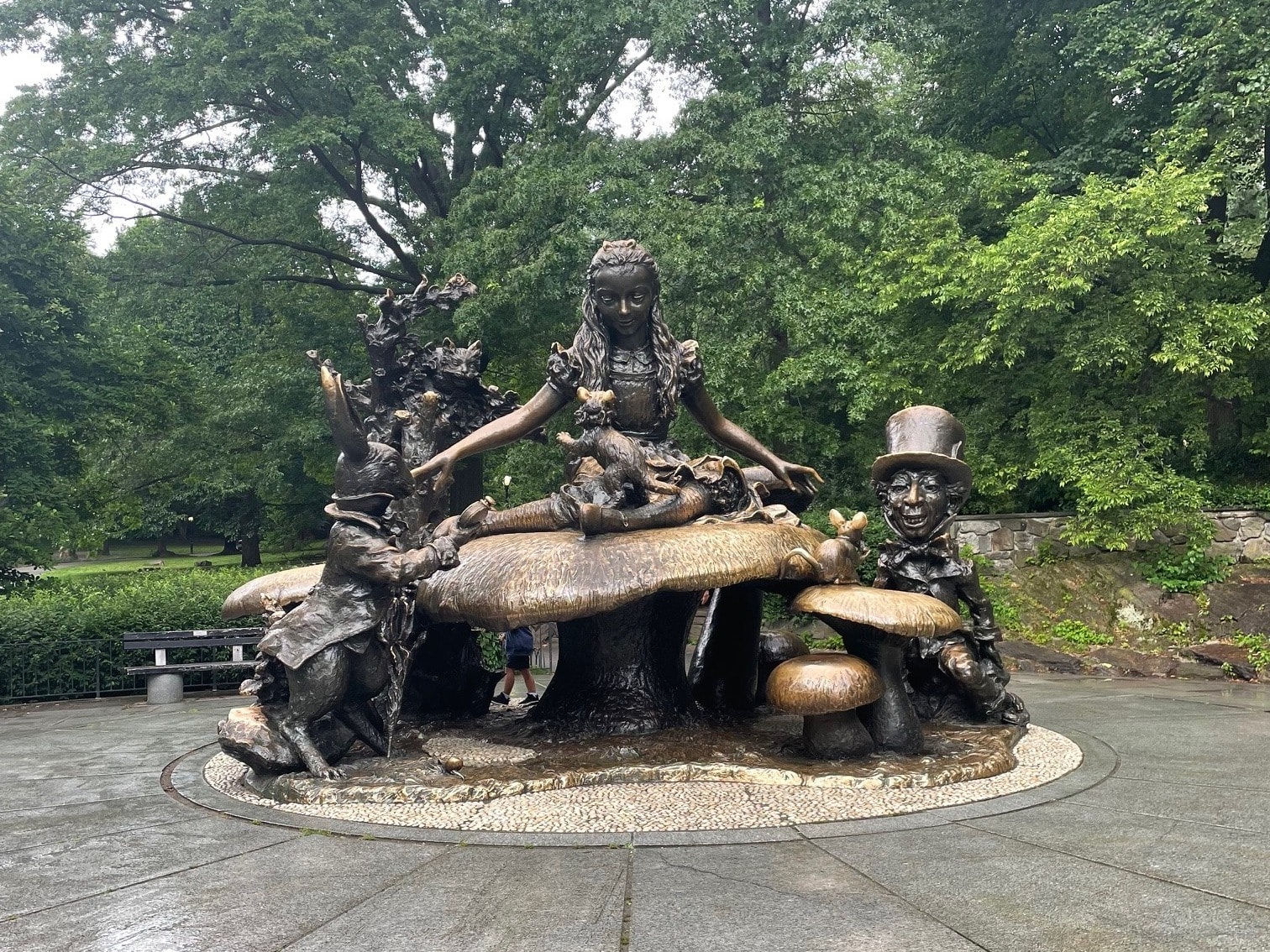 The Alice in Wonderland bronze sculpture set in a New York City park surrounded by greenery on a cloudy day. 