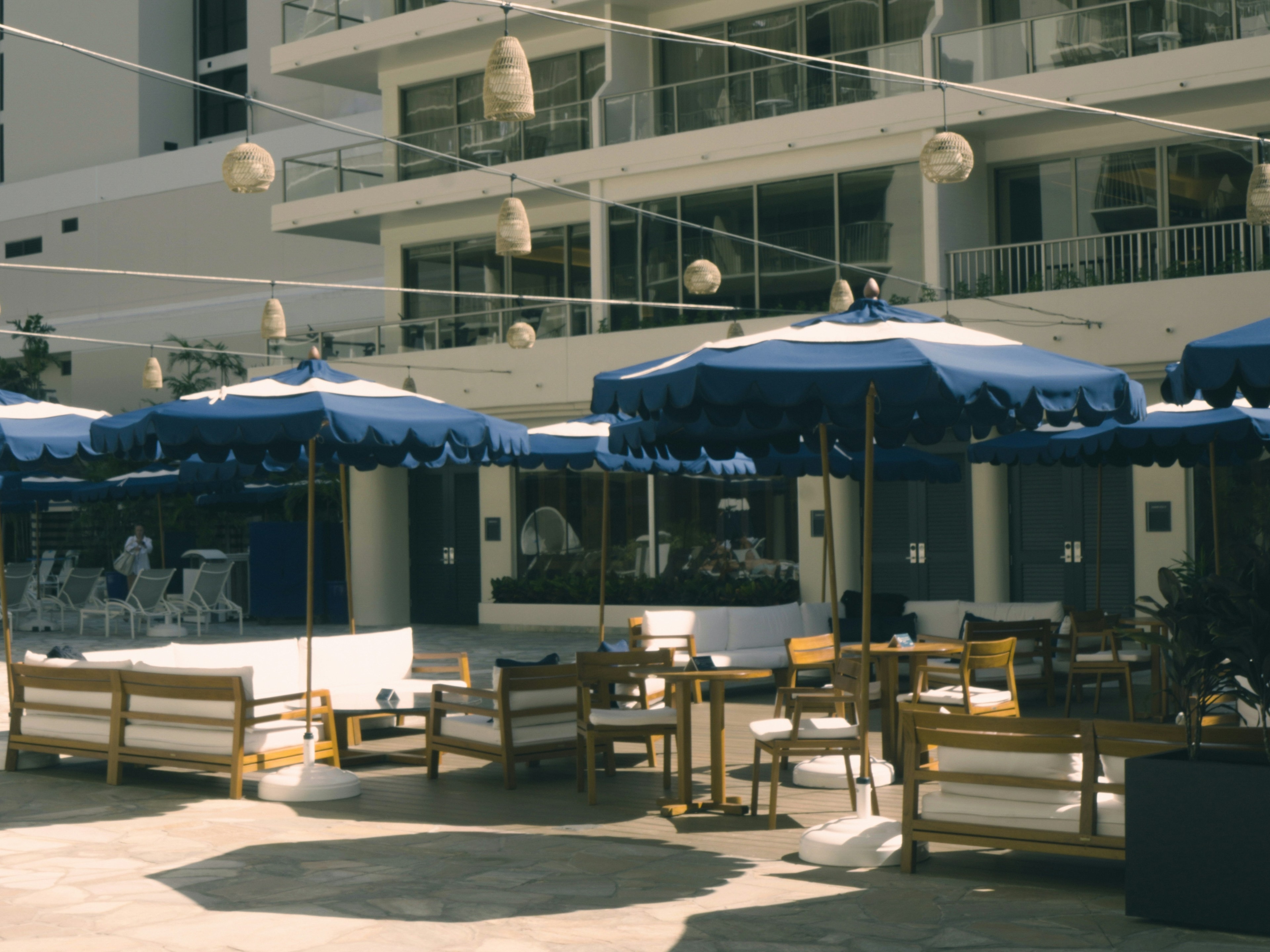 An outdoor seating area with blue umbrellas and wooden benches, is set against a multi-story building backdrop.