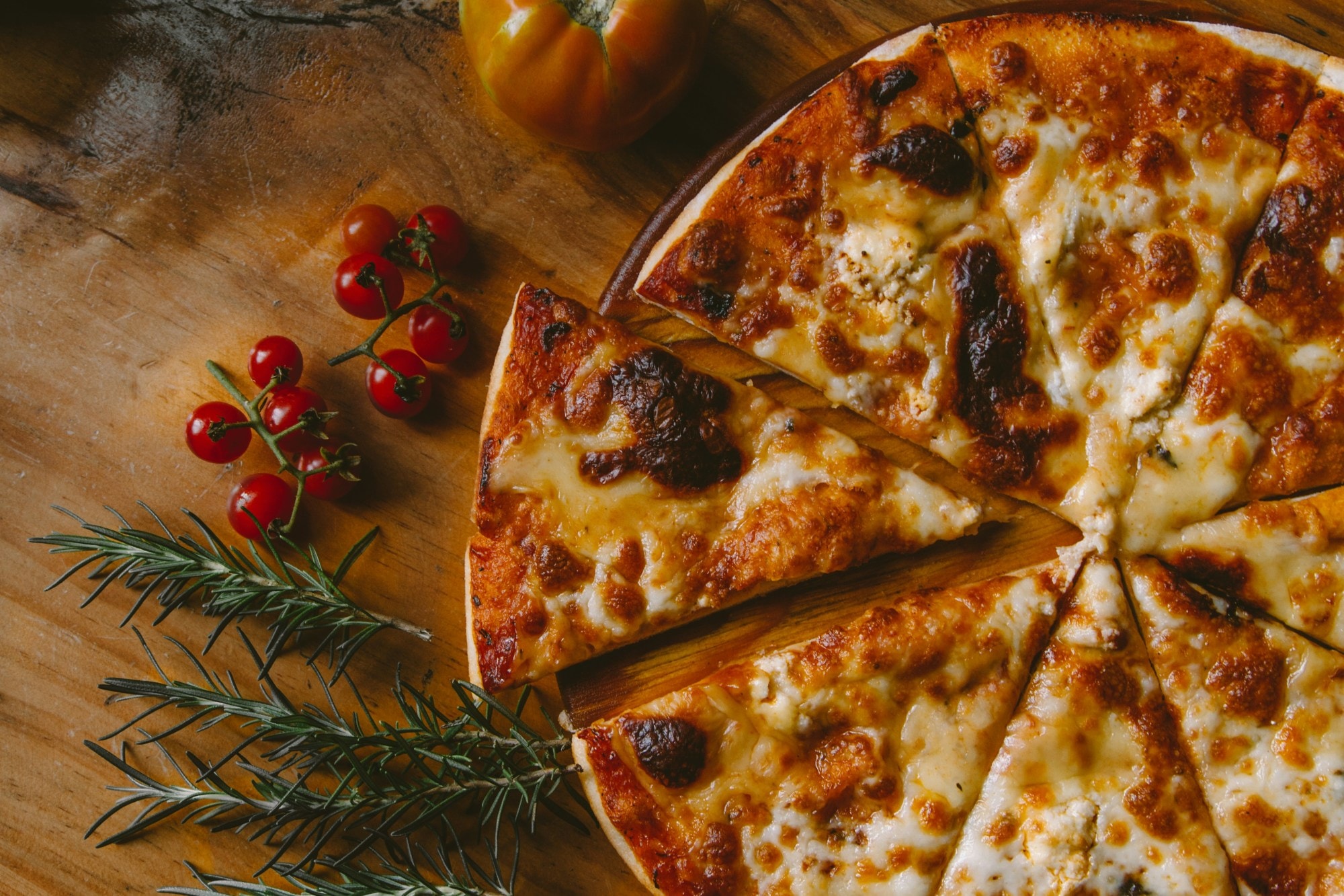 Sliced cheese pizza arranged on a cutting board amid cherry tomatoes and sprigs of rosemary. 