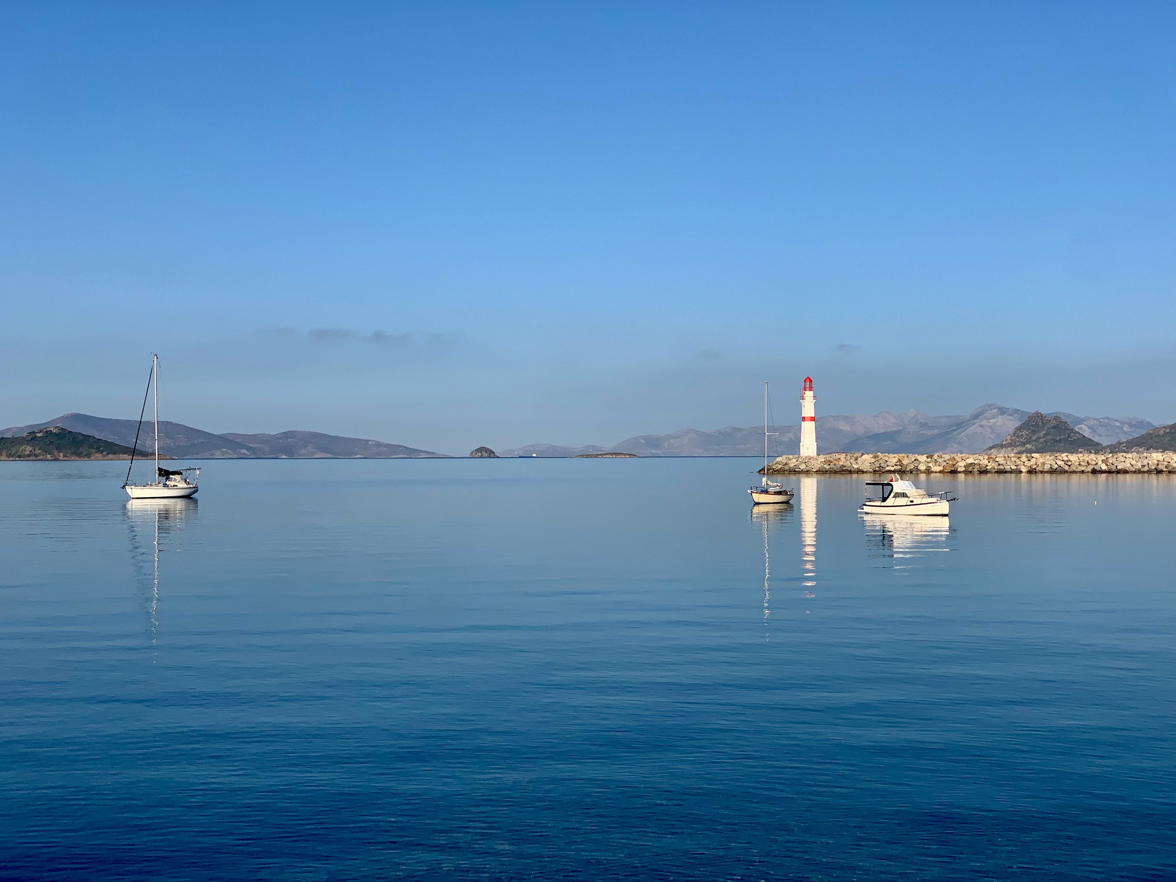 A serene water scene with a few boats and a lighthouse flanked by low hills.