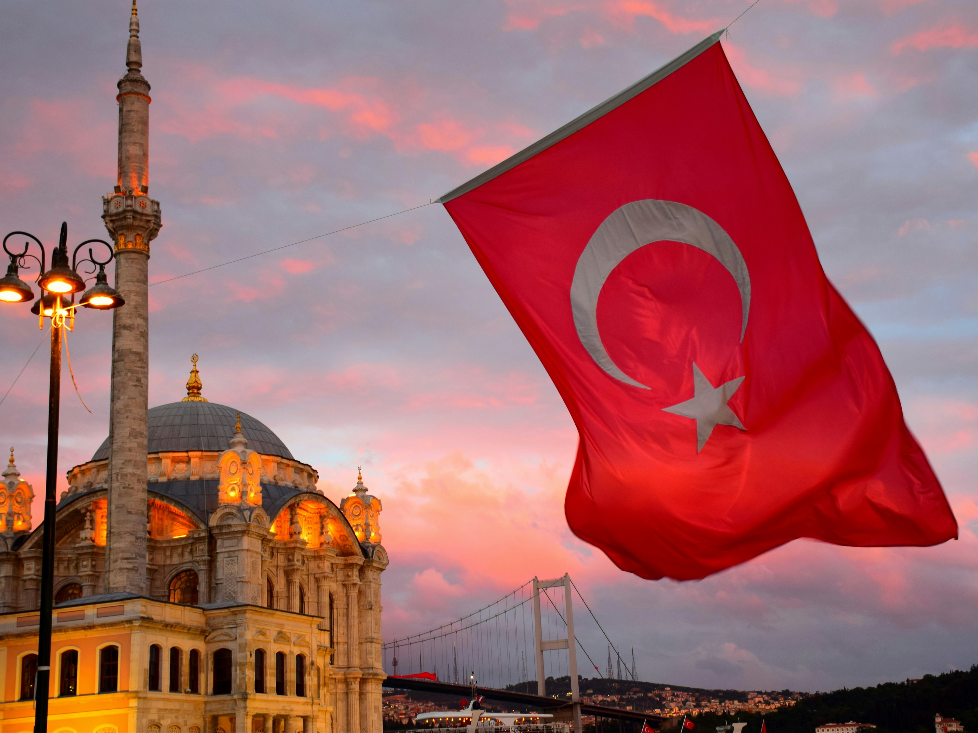 A twilight scene in Turkey, with a majestic mosque and bridge backdrop, with a large Turkish flag in the foreground.